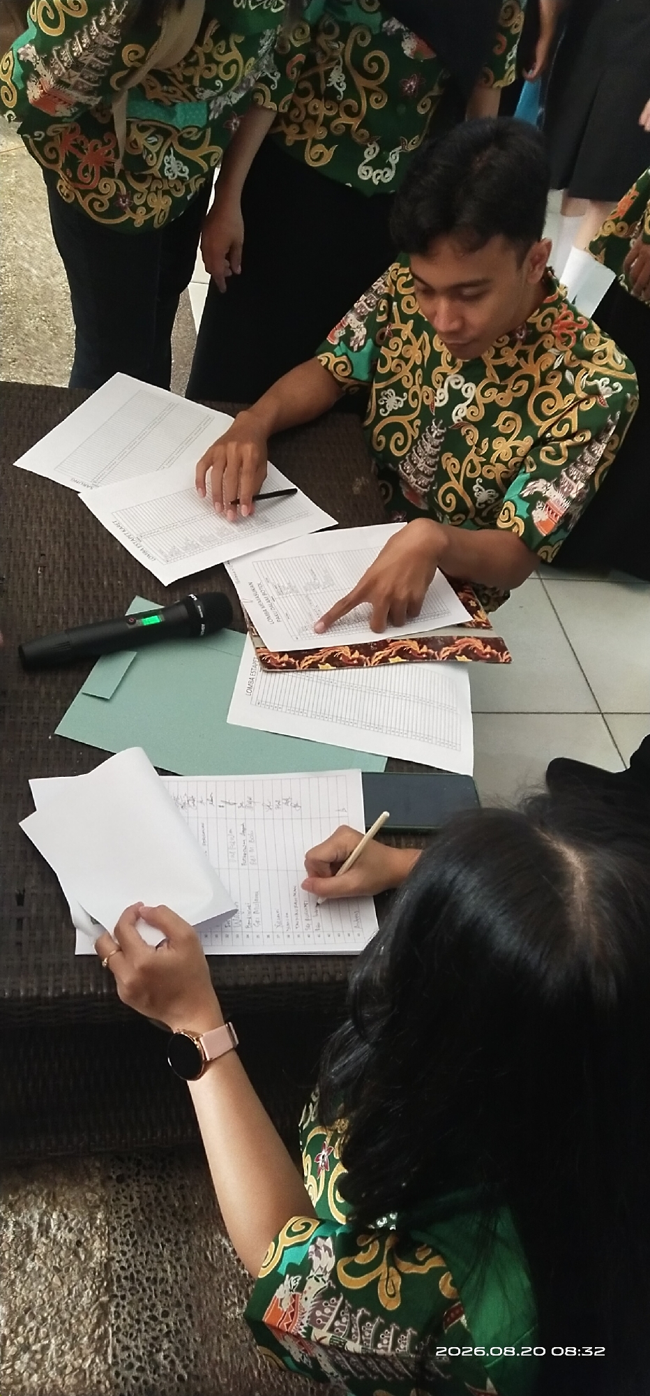 In Tamiang Layang, Indonesia, two individuals are engaged in an activity at a table, likely a desk. A young man, wearing a green and gold patterned batik shirt, is seen looking down at a document with his left hand resting on it. His right index finger is extended, pointing towards the paper. Beside him, another person, mostly obscured from view, is also looking at papers. A black microphone with a green light is placed on the table to the left of the papers. To the right, a person with long dark hair is writing on a sheet of paper with a pencil, holding a piece of paper in their left hand. They are wearing a pink watch on their left wrist and a similar green and gold batik shirt as the young man. The table surface appears to be a woven material, possibly rattan. The background is blurred, but suggests an indoor setting. A timestamp on the bottom right of the image reads "2026.08.20 08:32".