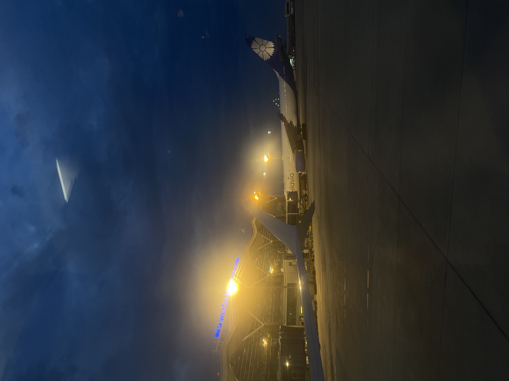 The image presents an illuminated airport scene at night or early morning, viewed from inside a passenger airplane parked on the tarmac. The sky is a deep, dark blue, suggesting the absence of daylight.
In the foreground and mid-ground, the wing of a large passenger jet is prominently visible, extending from the lower left towards the center. The aircraft's tail, seen in the upper right, features a distinctive white stylized flower or sunburst logo on a dark blue background, characteristic of Belavia airlines. Partially visible text on the fuselage reads "elavia," further confirming the airline.
Beyond the aircraft's wing, a modern airport terminal building is brightly lit by numerous yellow and white artificial lights, creating a warm glow and some lens flare effects in the photograph. Blue illuminated text on the terminal building clearly displays "NHÀ GA QUỐC TẾ CAM R..," which translates to "CAM RANH INTERNATIONAL TERMINAL," confirming the location as Cam Ranh International Airport in Bac Cam Ranh Ward, Vietnam.
The tarmac below is dark and appears paved, with subtle lines visible. No people are discernible in the frame. The scene captures a quiet moment at the airport, with the aircraft likely parked at a gate, awaiting boarding or preparing for departure. The overall atmosphere is still and calm, typical of an airport during the late hours or very early morning.