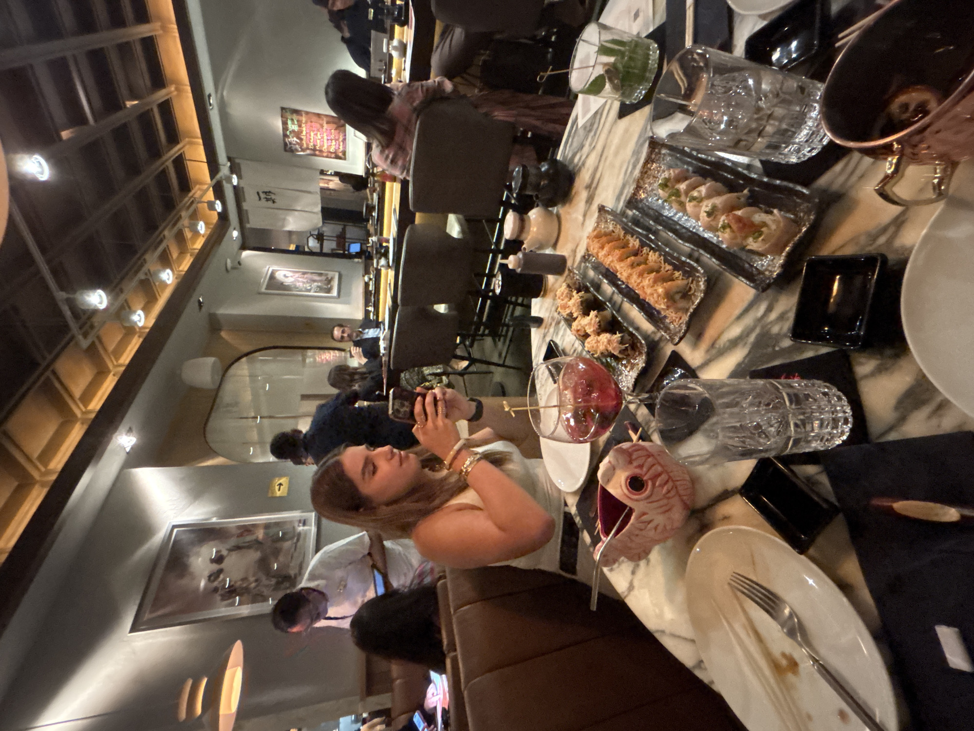 The image captures an indoor scene within a modern Japanese or Asian fusion restaurant in Mexico City, Mexico, during what appears to be evening hours due to the warm, artificial lighting. The perspective is from slightly above and to the left of a long counter or communal table.
In the foreground, a young woman with long brown hair, wearing a light-colored top and a gold bracelet, is actively holding a smartphone, seemingly capturing a photo or video of the table's offerings. In front of her, various dishes are laid out on a light-colored marble-patterned table with dark placemats. These include plates of sushi rolls, some topped with what appears to be crunchy tempura flakes, and other fried appetizers. A red cocktail in a stemmed glass, a clear glass with ice, and a distinctive pink fish-shaped ceramic cup with a straw are also visible on the table. An empty white plate with a fork rests to the right.
Behind this woman, other patrons are seated at similar tables or a bar, some engaged in conversation. One woman with dark hair is visible from the back. Further in the background, several individuals are seated, including a man in a light blue shirt. The restaurant features a stylish interior with a high, wooden slatted ceiling incorporating warm recessed lighting. Framed artwork adorns the walls, including a black and white image and a more colorful piece. A traditional Japanese *noren* (fabric curtain) with vertical Japanese characters, possibly "一番" (Ichiban), hangs in the mid-ground, suggesting a culinary theme. The atmosphere appears lively and upscale casual.