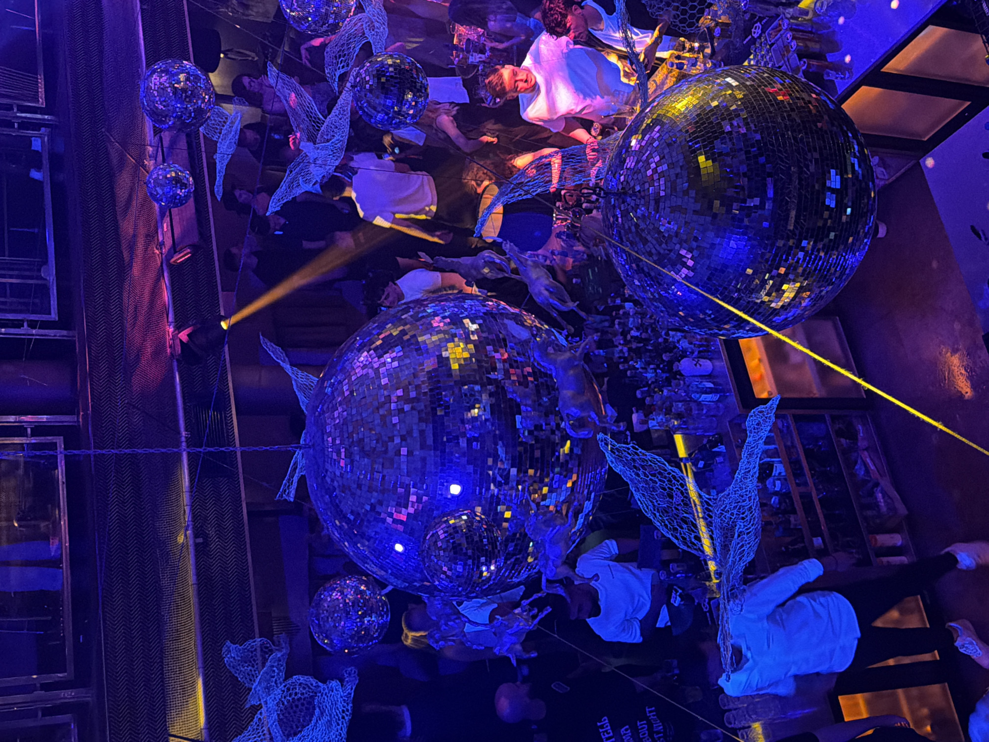 The image captures a vibrant, dimly lit nightclub interior, likely in Sofia, Bulgaria, with a focus on multiple large, sparkling disco balls suspended from the ceiling, casting fragmented light across the scene. The atmosphere is energetic, with a crowd of people visible, many appearing to be socializing and dancing.
In the foreground, a man in a light blue shirt and dark pants stands near a bar area, which is stocked with bottles. To his right, another man in a white t-shirt is engaged in conversation with someone out of frame. The background reveals more patrons, some interacting, others dancing, all bathed in the multicolored glow of the lighting and disco balls. The decor appears modern and industrial, with exposed ceilings and metallic elements. The overall impression is a lively night out, characteristic of a popular nightclub venue.