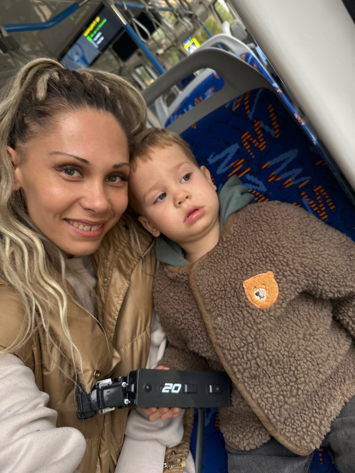 A woman with blonde hair pulled into a ponytail and a young child are seated next to each other on a bus, likely in or near Rusanovo, Russia. The woman is smiling at the camera, holding a small black device with a digital display showing "20". She is wearing a tan puffer jacket over a cream-colored top. The child, also looking towards the camera but with a slightly pensive expression, is wearing a brown, fuzzy jacket with a bear patch on the chest and a green hooded sweatshirt underneath. The bus seats are blue with a pattern of orange and yellow zigzags. Overhead, a digital display panel shows green and yellow text, likely indicating route information. The overall environment suggests an indoor setting on public transportation.