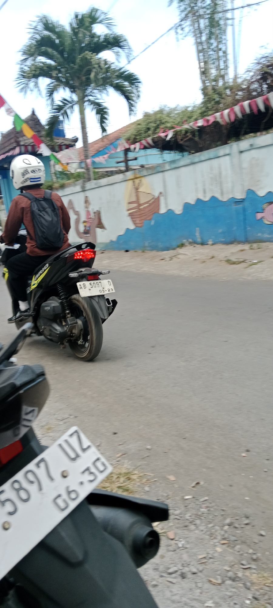 An over-the-shoulder shot captures a person riding a black motorcycle with a brown long-sleeved shirt and a black backpack. The motorcycle's license plate reads "AB 5097 D 03.29". In the foreground, another motorcycle's license plate is visible, displaying "5897 UZ 06.30". The background features a light blue wall with a mural depicting a ship and wavy blue lines, and a palm tree. Decorative flags are strung across the top of the wall. The scene appears to be on a paved road, consistent with the village of Purwomartani, Indonesia, given the style of the motorcycles and the surrounding environment.