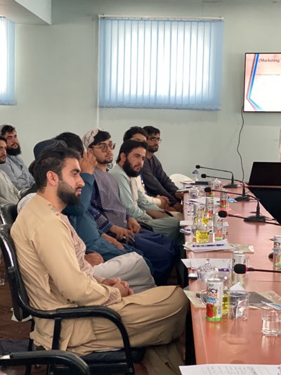 A group of men sit in a conference room. The room is equipped with a projection screen displaying a presentation slide, microphones, and bottles of juice on the table. The men appear to be attending a meeting or lecture. The location is likely Shurandam, Afghanistan.
