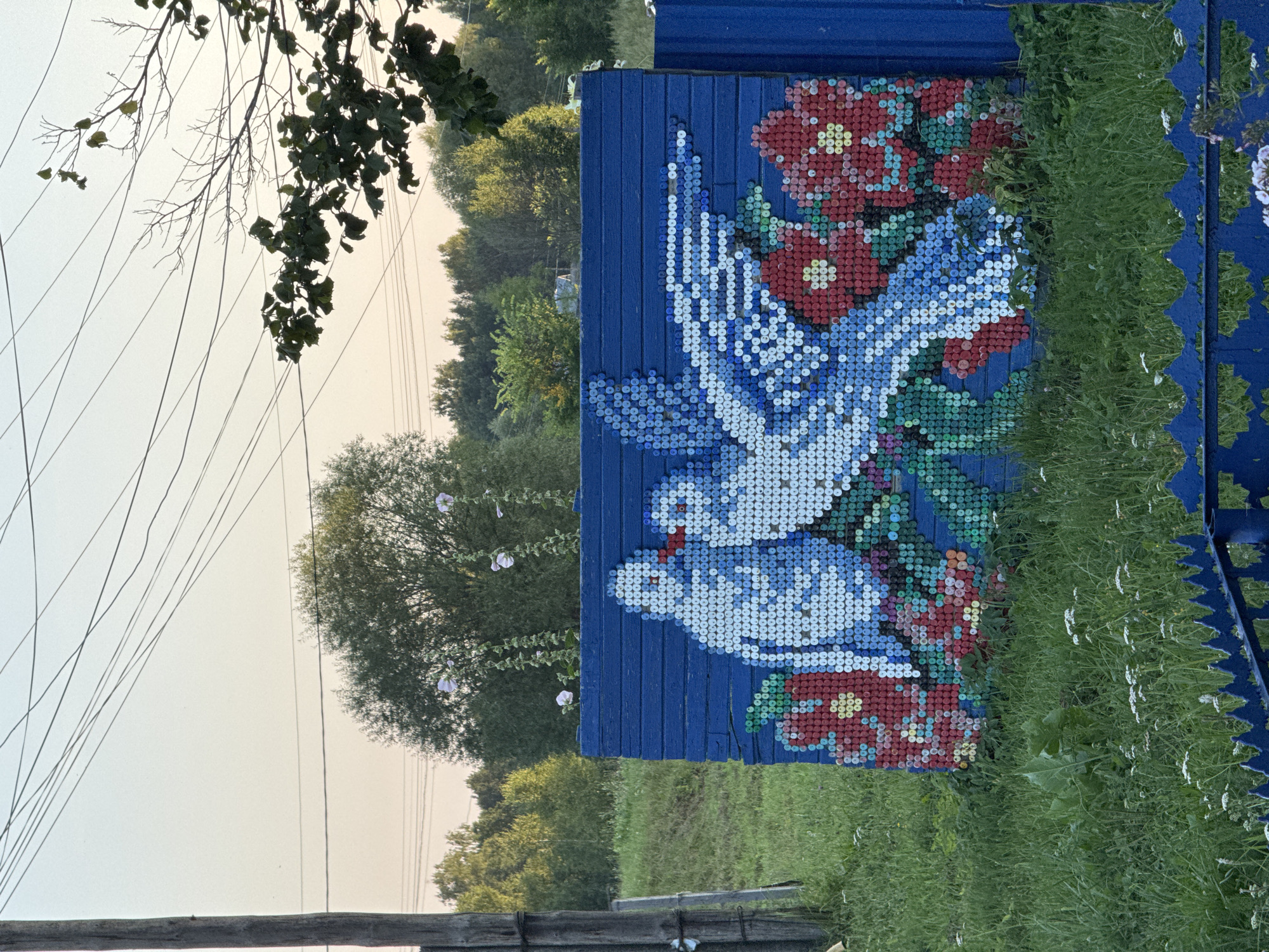 This image depicts an outdoor scene in the city of Lysaya Gora, Russia, featuring a colorful mosaic artwork on a blue wooden fence. The mosaic, made of bottle caps, depicts two white doves with blue accents on their wings, facing each other, with red beaks touching. They are positioned above a cluster of red flowers with green centers and stems, and some green foliage. The artwork is located on a blue, horizontally planked fence. To the left of the fence, a wooden utility pole with wires running overhead is visible. The background consists of lush green grass, scattered small white wildflowers, and a dense line of trees under a clear, pale sky. The overall impression is one of folk art decoration in a rural or semi-rural setting. There are no people visible in the image.