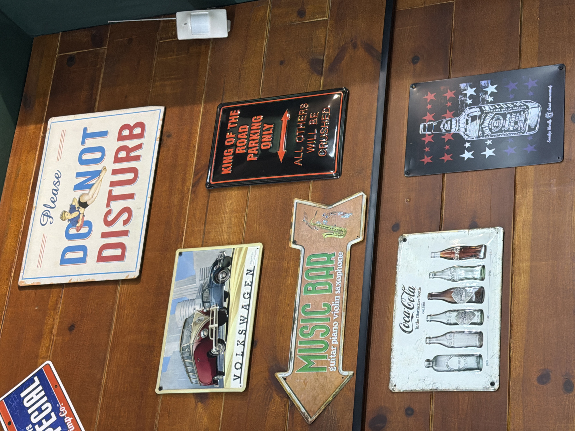 A collection of decorative signs hangs on a wooden wall. The signs feature various images, including a woman in a swimsuit, Volkswagen cars, musical instruments with the words "MUSIC BAR", different Coca-Cola bottles, and a bottle of Jack Daniel's Tennessee Whiskey surrounded by stars. One sign reads "KING OF THE ROAD PARKING ONLY. ALL OTHERS WILL BE CRUSHED." A motion sensor is located in the upper-left portion of the frame. There is no indication of location in the city of Budva, Montenegro, or any people present.