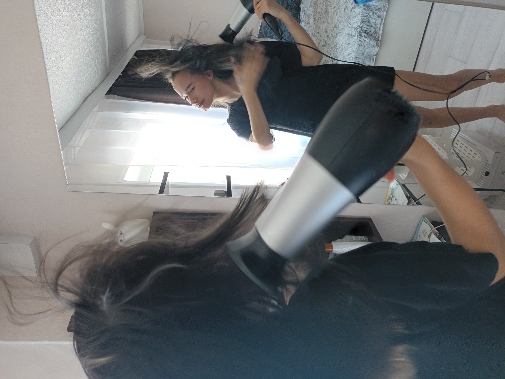 A person is using a hairdryer to dry their hair in a room with a mirror. The reflection shows the person in a black top and a bed with patterned bedding. There is a window with white curtains. The location is indicated as Ольховка, Russia.