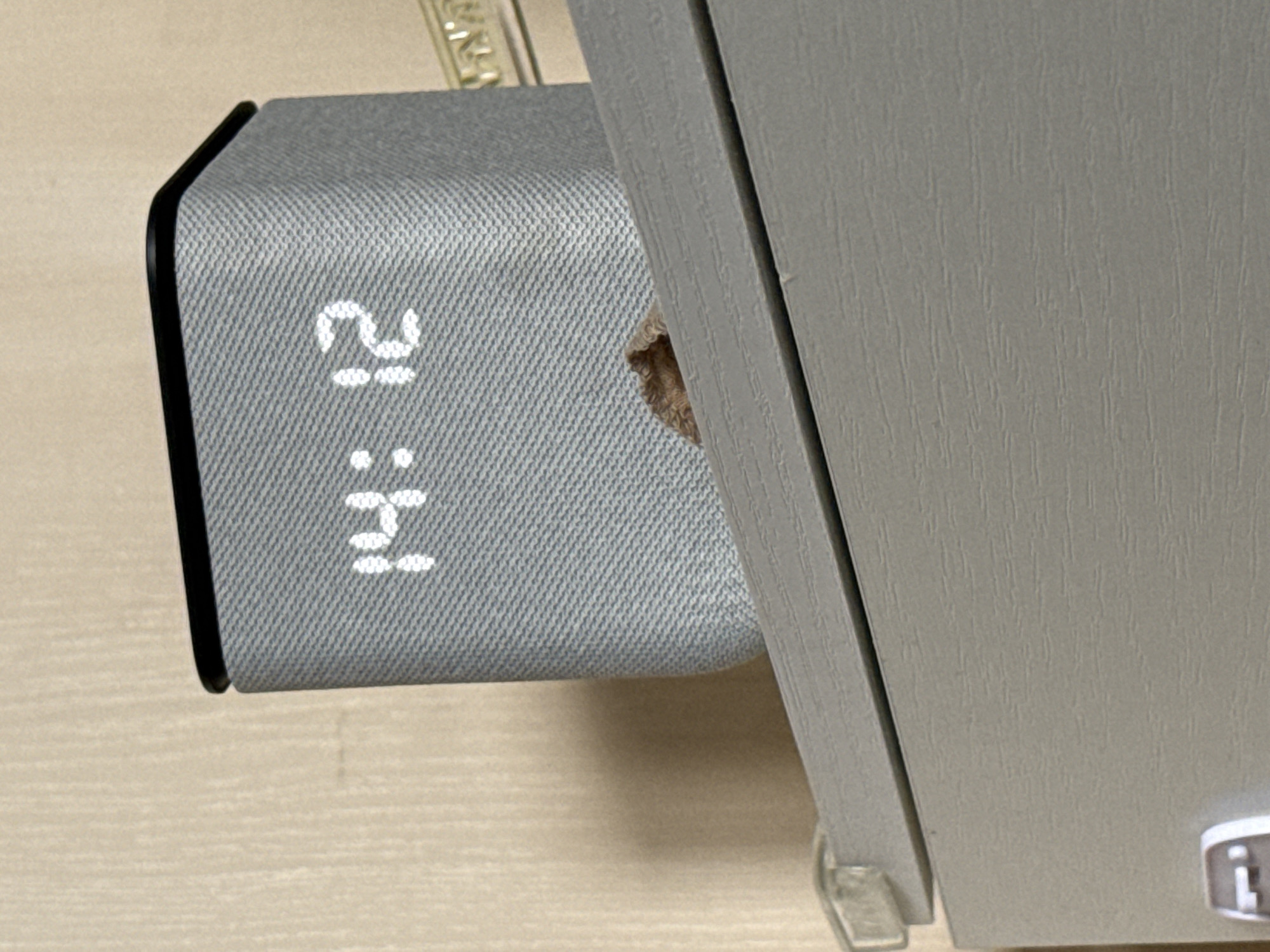 The image shows a digital clock displaying 14:21, positioned on a table next to a light gray cabinet. The clock has a fabric-covered exterior. A small, brown object is visible in the space between the clock and the cabinet. The environment appears to be indoors, possibly a home or office. The location in the city of Yakutsk, Russia cannot be confirmed from the image.