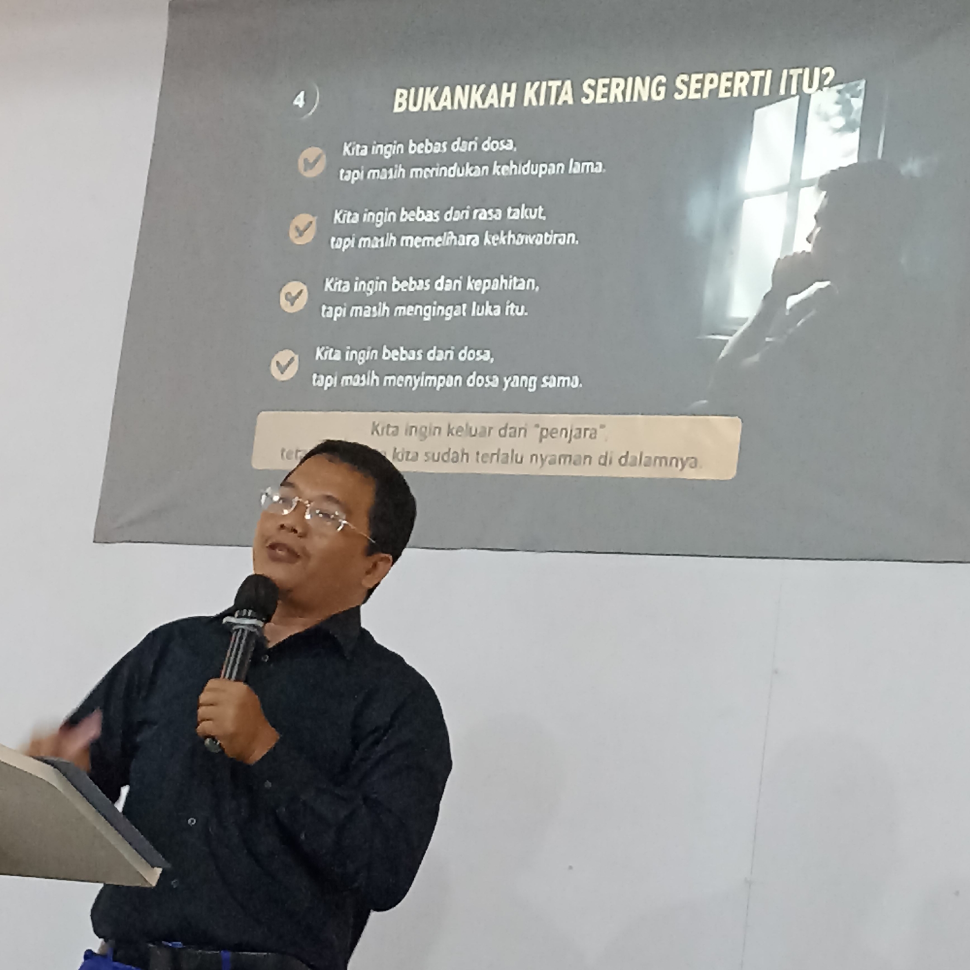 A man wearing glasses and a black collared shirt is speaking into a microphone. He is standing in front of a projector screen that displays text in Indonesian. The screen has a title that reads "BUKANKAH KITA SERING SEPERTI ITU?" (Aren't we often like that?) followed by a list of statements with checkmarks next to them. The man's left hand is holding the microphone, and his right hand is gesturing. Behind the screen, a silhouette of a person is visible, looking out of a window. The background appears to be a wall, and the lighting suggests an indoor setting. The image was taken in Magelang, Indonesia.