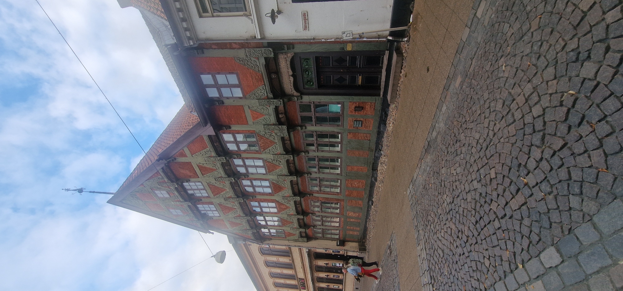 The image depicts a cobblestone street in front of a historic, multi-story building with red brick and green timber framing, located in Kolding, Denmark. The building features numerous windows, decorative geometric patterns on its facade, and a prominent gable. A dark wooden double door is visible on the ground floor. To the right of this building, a white facade with a street sign reading "Torvegade" is partially visible, indicating the street name. Two individuals are walking on the sidewalk in the foreground. The sky above is partly cloudy. The overall scene suggests a well-preserved historic urban environment.
