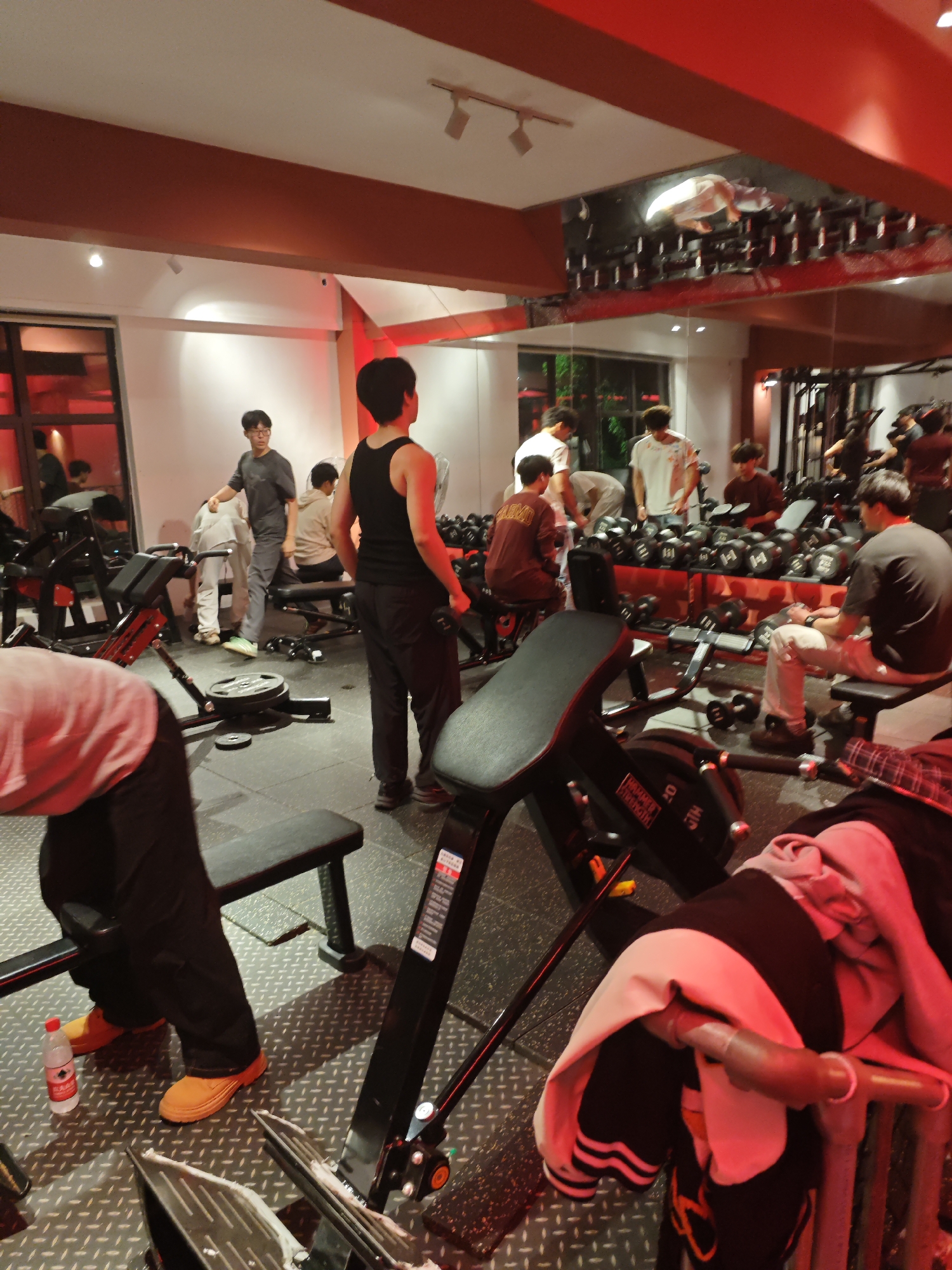 The image shows a busy gym interior in Chongqing High-tech Zone, China. Several people are engaged in various exercises. There are multiple weight benches and dumbbell racks. One man is standing in a tank top, seemingly between sets. Another man is seated, resting between reps. Others are near the dumbbell rack, likely selecting weights. A water bottle rests near a pair of orange work boots in the foreground. The room has a reflective ceiling with racks of dumbbells visible.