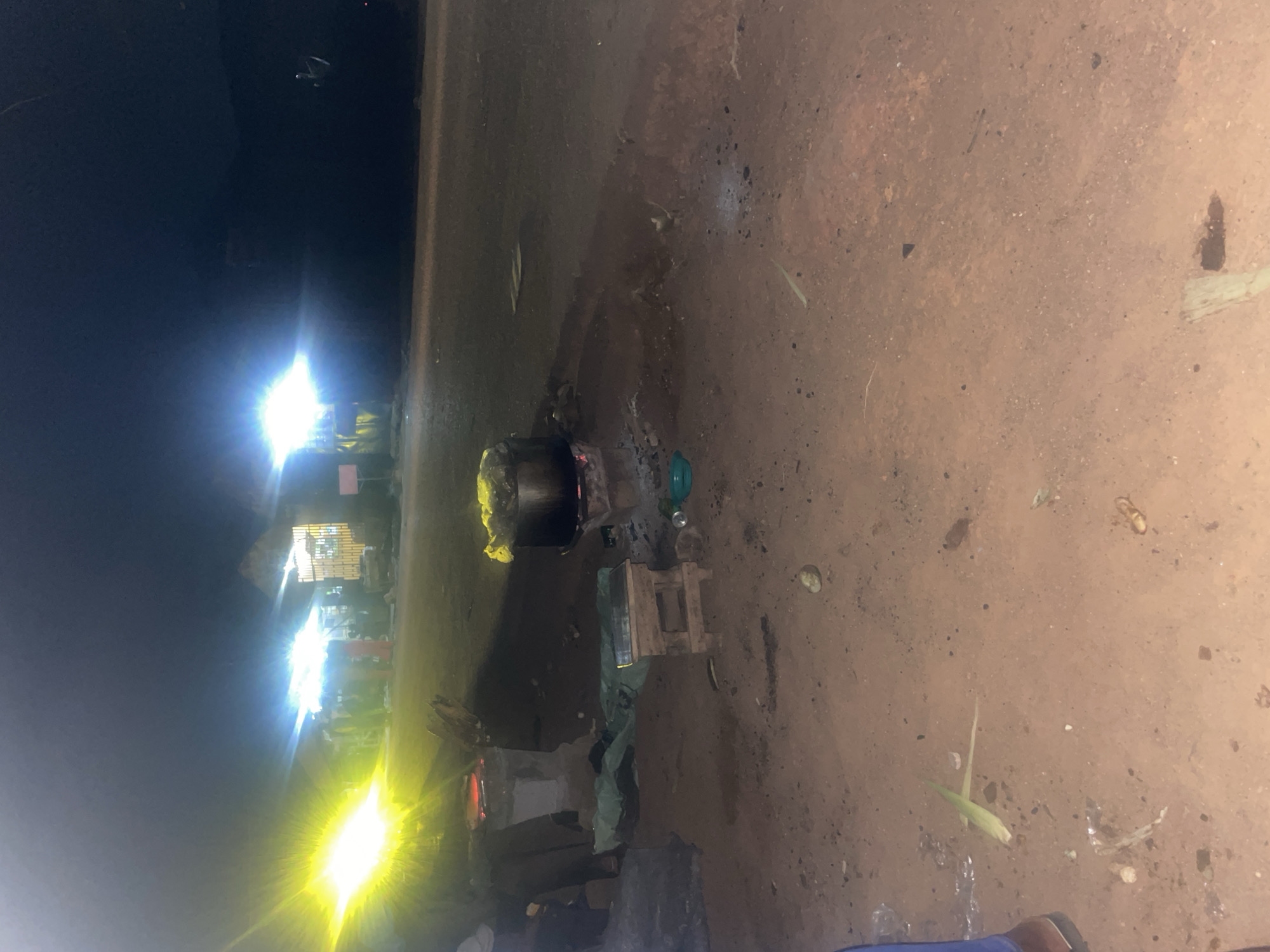 The image depicts an outdoor scene at night, likely in Du, Nigeria. Bright artificial lights, some appearing to be from streetlights or stalls, illuminate the area, casting strong glows and creating lens flares. In the center of the frame, a large pot sits over what appears to be a fire, suggesting cooking is taking place. Around the pot, there are various objects, including what looks like a wooden stool or stand, a green container, and some discarded materials. In the background, there are glimpses of structures that resemble market stalls or simple buildings, also lit by artificial lights. The ground is uneven and appears to be dirt or a similar natural surface, with some scattered debris and vegetation. A person's legs and feet, wearing dark footwear, are visible in the lower right corner, indicating a human presence. The overall impression is of a bustling, albeit dimly lit, nighttime environment, likely a public space where food is being prepared or sold. The time of day is clearly night due to the artificial lighting and dark sky. There are no discernible weather conditions visible, but the ground appears slightly damp in places. There is no visible text in the image.
