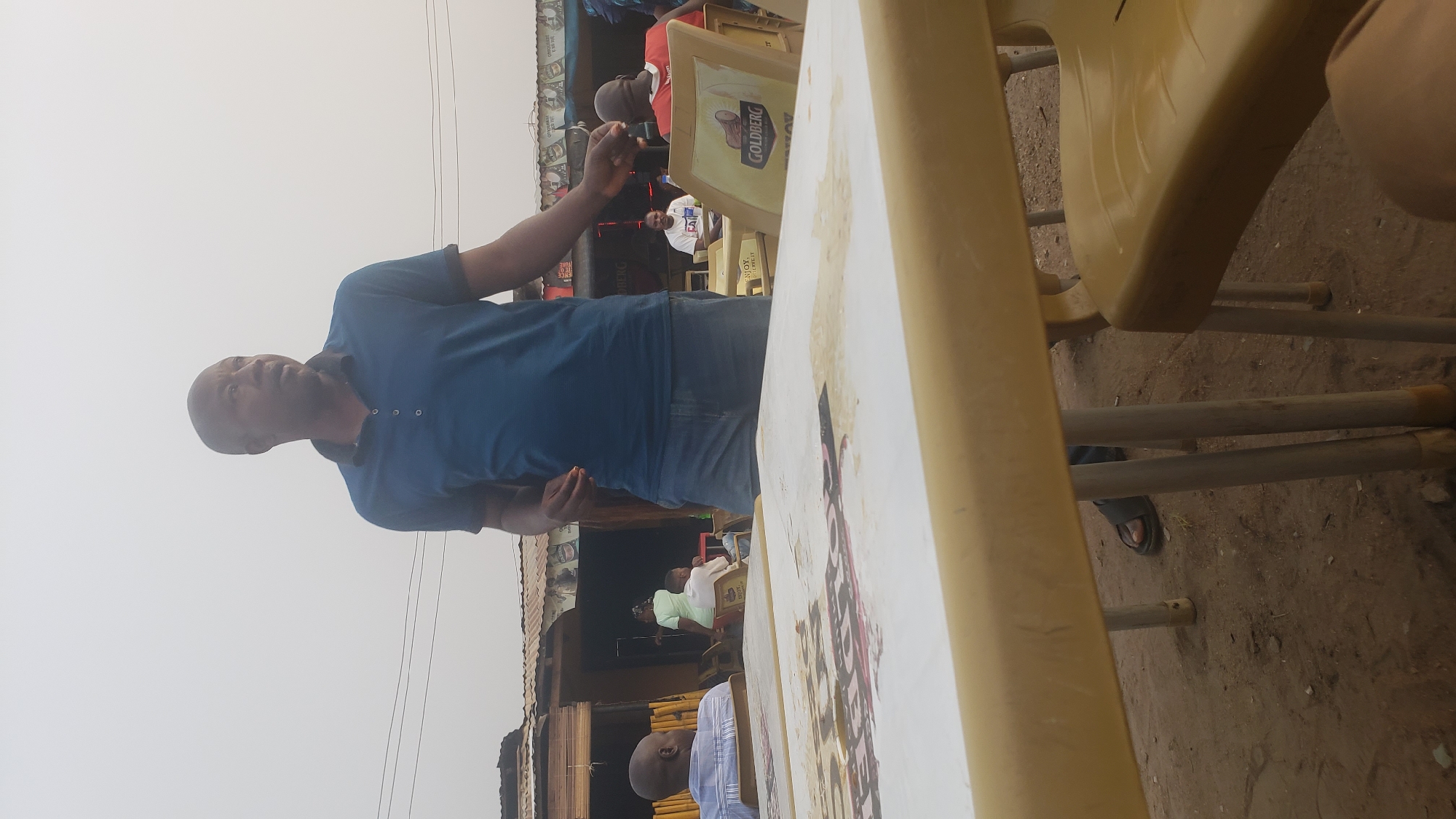 A medium, horizontally-oriented outdoor shot depicts an adult Black male standing near a plastic table and chairs in what appears to be a public space, potentially a roadside establishment, in Narayi, Nigeria. The man is facing towards the left side of the frame with his body oriented towards the camera; he is wearing a short-sleeved, collared blue shirt and jeans. He has short hair or is bald.
In the foreground, a cream-colored plastic table dominates the view, partially obscuring the scene behind it. To the right of the table are yellow plastic chairs, one of which is partially occupied by an unseen person, evidenced by an exposed leg.
Behind the man and table, a series of low-lying buildings with corrugated metal roofs and storefronts create a backdrop. In this area, at least three other people can be discerned. One is wearing a white t-shirt and seated, the others have partial bodies visible.
The ground beneath the tables and chairs appears to be bare earth. Overhead, telephone wires or electrical cables are visible. The sky is pale, suggesting either overcast conditions or strong sunlight. The setting appears to be modest and functional, possibly a small business or local gathering spot.