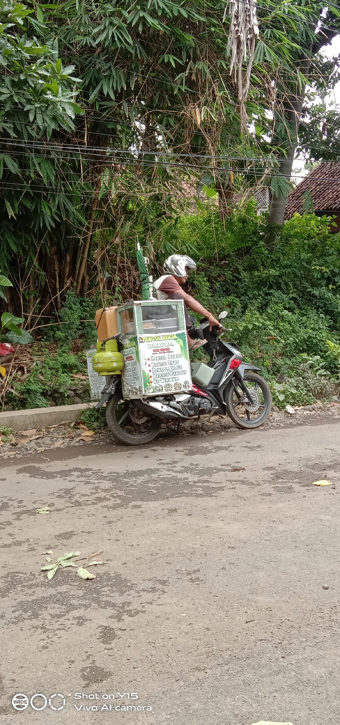 A street vendor on a motorcycle with an attached food cart is parked on a paved road next to dense green foliage in Brabe, Indonesia. The vendor, wearing a white and black helmet and a brown shirt, is seated on the dark-colored motorcycle, facing right.
The green and white food cart features a glass display. A green gas cylinder is mounted at the rear of the motorcycle, supporting the cart, and a green umbrella is also attached to the structure. Text on the cart advertises "PENTOL BERKAH JAYA" and lists items such as "PENTOL BAXBO," "PENTOL TARU," "PENTOL KASUT," "KERIRIK PANDOT," and "ES WAWAN," likely referring to different types of Indonesian meatballs and a drink.
The background is dominated by lush green bamboo and other tropical vegetation, suggesting a rural or semi-rural setting. In the upper right, a section of a reddish-brown tiled roof of a building can be seen. The asphalt road in the foreground is visible, showing some wear and scattered dry leaves. The scene is brightly lit, indicating daytime. A watermark "Shot on Y15 Vivo AI camera" is present in the lower left corner.