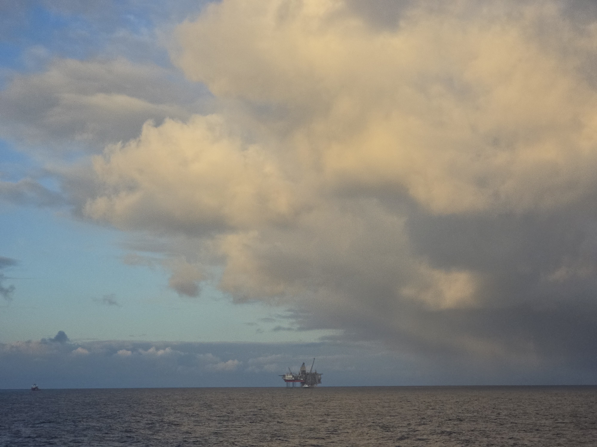 A wide shot depicts an oil rig in the middle of the ocean under a cloudy sky. The sky transitions from blue on the left to gray and white clouds on the right, with some clouds tinged with yellow. The ocean is dark and calm. A small vessel is visible in the distance to the left of the oil rig.