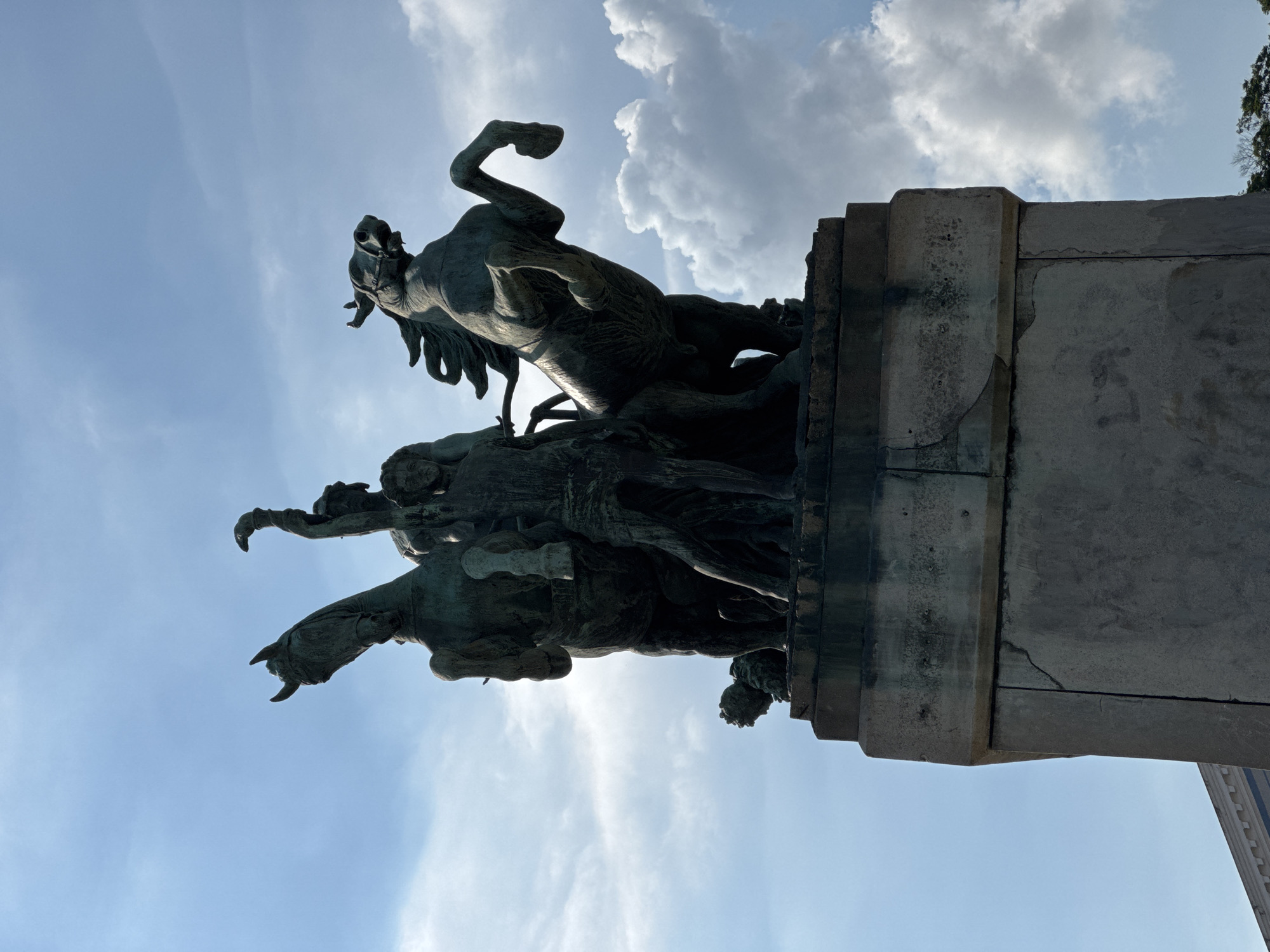 This is a low-angle, medium shot of a bronze equestrian statue situated on a stone pedestal against a bright, partly cloudy blue sky. The statue depicts two horses rearing up, with a male figure standing between them, holding what appears to be a rein or whip aloft. Another figure, possibly also male, is partially visible behind the main figure. The stone pedestal shows signs of wear and cracking. The statue is located in the city of Niterói, Brazil, specifically at the Parque da Cidade, a prominent viewpoint overlooking the Guanabara Bay and Rio de Janeiro. The time of day appears to be daytime, judging by the bright sky and diffused sunlight. The overall scene is monumental and somewhat dramatic due to the rearing horses and the upward angle of the shot.