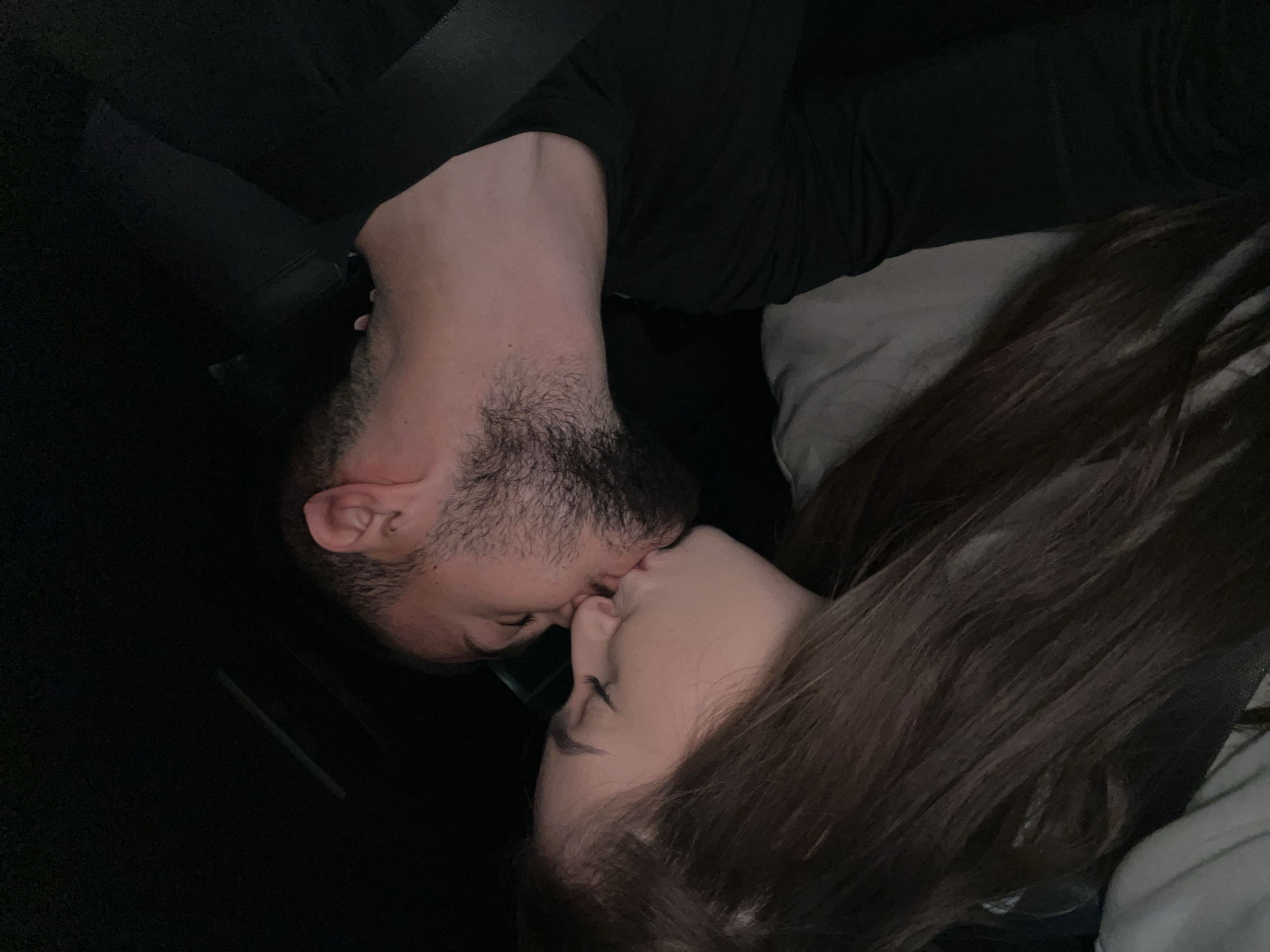 The image captures a close-up, intimate moment between a man and a woman kissing. The man, positioned on the left side of the frame, has a beard and stubble and is wearing a dark, long-sleeved top. A dark seatbelt is visible across his shoulder and chest, strongly suggesting the setting is inside a vehicle, likely a car. The woman, on the right, has long, dark brown hair which partially obscures a light-colored pillow or cushion behind her head.
The lighting is dim, indicating that the photo was taken at night or in a dark interior. Their faces are in close profile, with lips touching, conveying a sense of deep affection and intimacy. The activity is a kiss between the two individuals. There are no visible location cues within the image itself to confirm the specified location of Plovdiv, Bulgaria, nor is any text visible. The overall mood is tender and private.