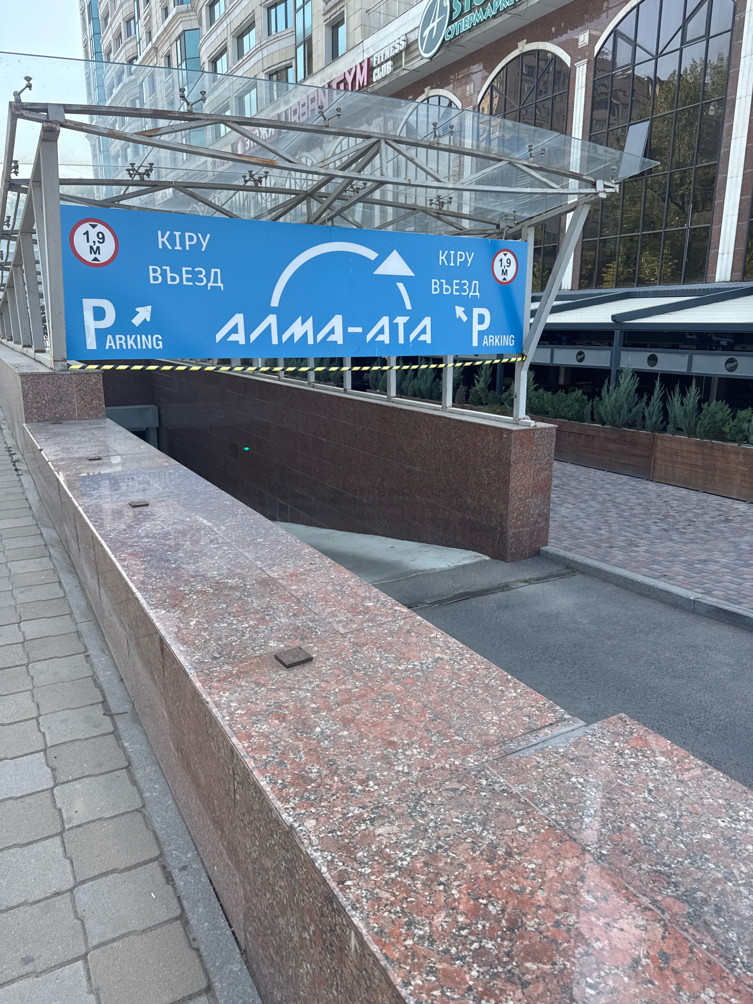 The image shows a parking entrance in Almaty, Kazakhstan. A blue sign with white lettering indicates "PARKING" and directional arrows. The sign also displays a height restriction of 1.9 meters. The entrance appears to be a ramp leading down into an underground parking area. The surrounding area is paved with stone and asphalt, with some planters containing evergreen shrubs. The building in the background features large arched windows. No people are visible in the image.