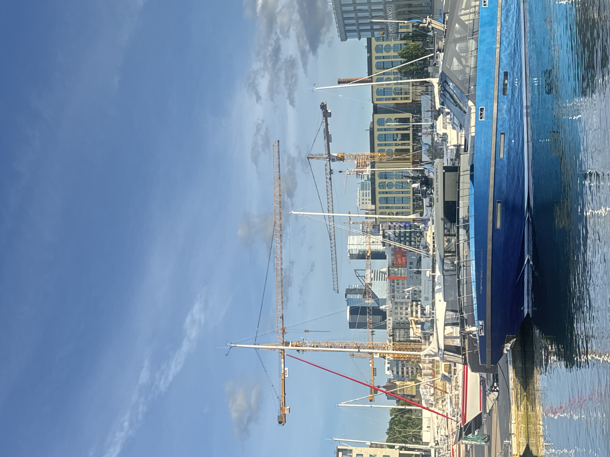 The image depicts a waterfront scene in Tallinn, Estonia, likely in the developing port area. In the foreground, several yachts are docked in a marina, with a prominent blue yacht in the center. Behind the yachts, a cityscape is visible, characterized by modern buildings and several construction cranes, suggesting ongoing urban development. The sky is a clear blue with scattered clouds. The water in the foreground reflects the surrounding structures and the sky. The overall atmosphere is one of a bustling port and developing urban environment.