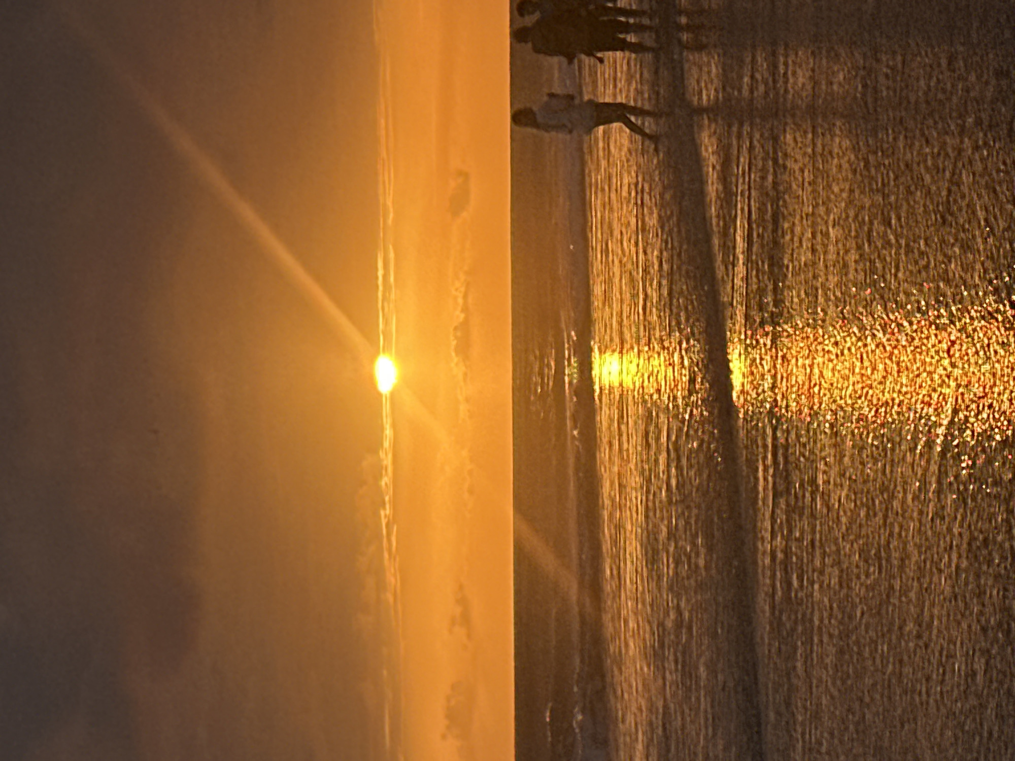 A serene sunset scene unfolds over the ocean in North Kuta, Indonesia. The sun, a brilliant orb, is positioned just above the horizon, casting a warm, golden-orange glow across the sky and the water. Wispy clouds are scattered across the upper portion of the sky, with a prominent ray of sunlight extending diagonally upwards. The ocean's surface is calm, reflecting the vibrant colors of the sunset, creating a shimmering path of light.
In the middle ground, several figures are visible standing in the shallow water near the shore. One figure, in the foreground on the right, appears to be a person walking away from the camera, their silhouette distinct against the bright water. Two other figures are seen behind and to the right of this individual, appearing as darker silhouettes. The waves are gentle, lapping at the shore and the feet of the people. The beach itself is not clearly visible, as the focus is on the water and the sunset. The overall impression is one of tranquility and natural beauty.