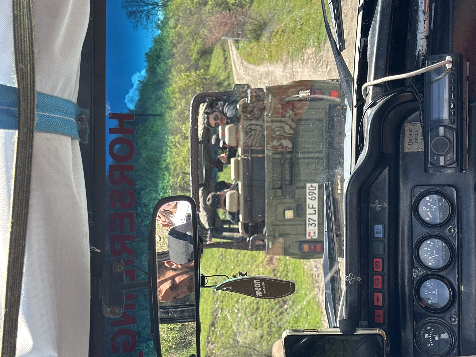 A vehicle in Haghartsin, Armenia follows another vehicle on a rural road. The interior of the vehicle includes a rear-view mirror with a reflection of the driver, a radio, an Areon air freshener, dashboard gauges, and a curtain. The license plate of the vehicle ahead reads "37 LF 690." The other vehicle is an open-top vehicle with people in it. The sky is visible through the front windshield.