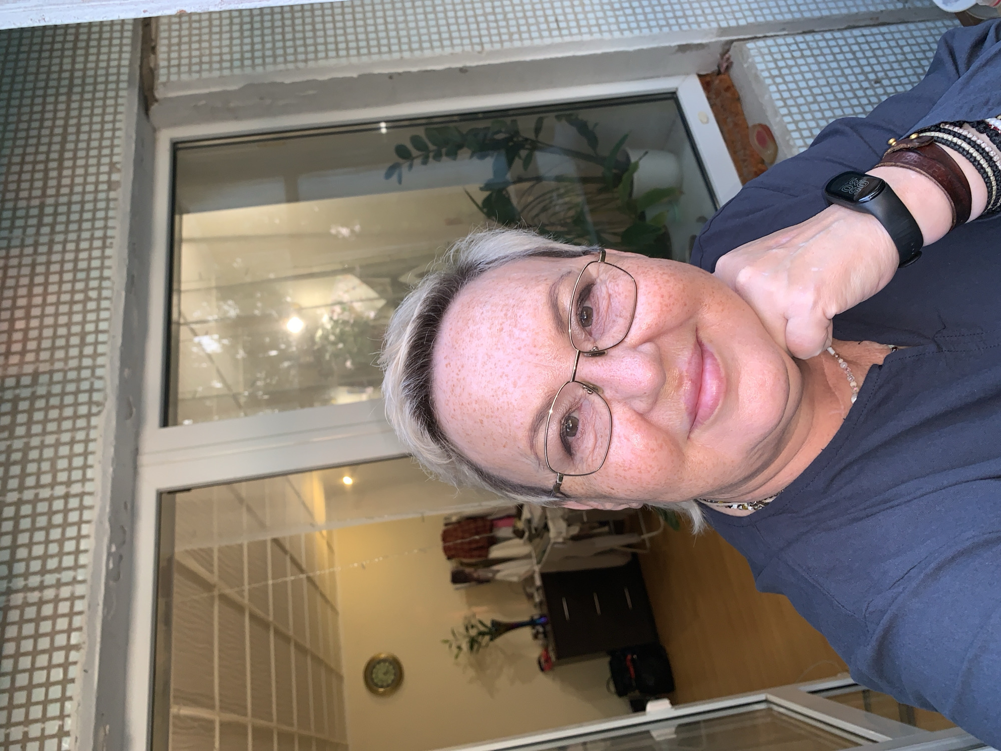 A medium shot, selfie-style portrait of a middle-aged Caucasian woman with blonde hair, freckles, and glasses. She is wearing a dark blue shirt and multiple bracelets on her left wrist, including a black smartwatch. Her right hand is resting on her chin. The background appears to be an interior space with a window. Visible elements include a ceiling with fluorescent lighting, a clock on the wall, a dresser with a vase of flowers, and some clothing hanging on a rack. The window shows a reflection of greenery and a potted plant. The overall setting suggests a home environment, possibly an apartment in Chaikovsky, Russia, given the architectural details and common interior design elements. The lighting suggests it is daytime.