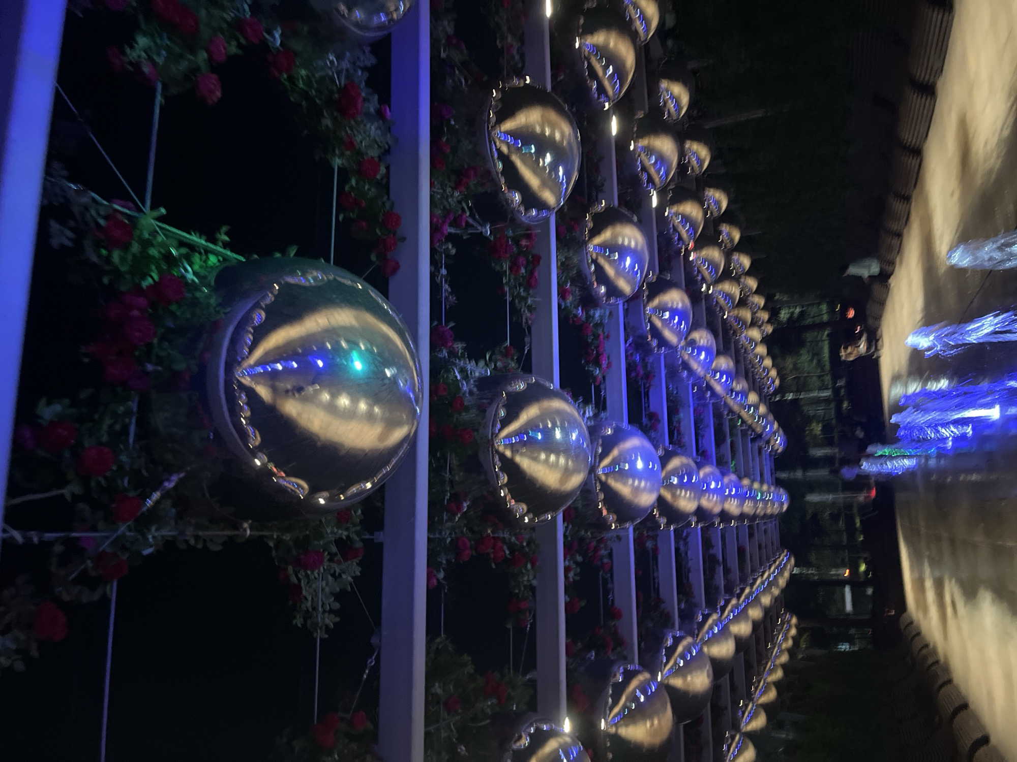 The image depicts an outdoor night scene, likely a public space with decorative elements. Overhead, rows of large, spherical, mirrored decorations are suspended, reflecting the ambient light and surrounding environment. These decorations are interspersed with artificial or real flowering vines, primarily red roses. Beneath the decorations, there's a walkway or patio area where water fountains are active, casting blue and green light onto the wet surface. Benches are visible along the sides of the walkway. Several indistinct human figures are present in the background, engaged in casual activity or simply present in the space. The overall impression is one of an illuminated, possibly festive or atmospheric, outdoor venue. The specific location in Krasnodar, Russia, is not identifiable from the visual information presented.