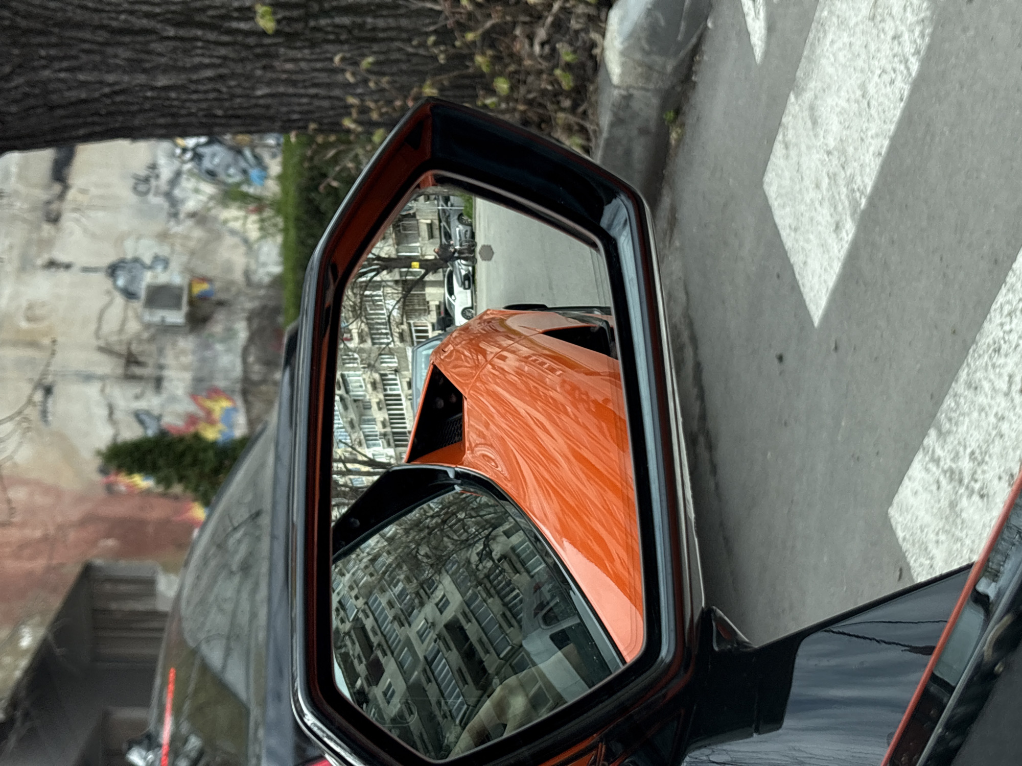 The image depicts the side mirror of a black car, reflecting an orange car, buildings, and surrounding urban elements. The buildings appear to be located in an urban area, likely in the city of Varna, Bulgaria. A tree is visible in the upper-left portion of the image. A zebra crossing is visible to the right of the car.