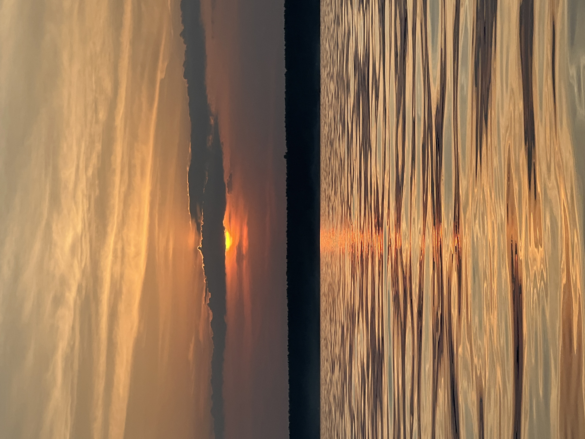 The image depicts a sunset over a body of water, likely a lake, with a dense tree line in the background. The sky is filled with warm hues of orange, yellow, and pink, with wispy clouds scattered across the horizon. The sun is partially obscured by a dark cloud, casting a brilliant orange glow on the water's surface. The water itself is calm, with gentle ripples reflecting the colors of the sky. The tree line appears dark and silhouetted against the vibrant sky, suggesting a forested area. The overall impression is one of tranquility and natural beauty. Based on the provided context of the image being from Borovoye, Russia, this scene captures the essence of its natural landscapes during sunset.