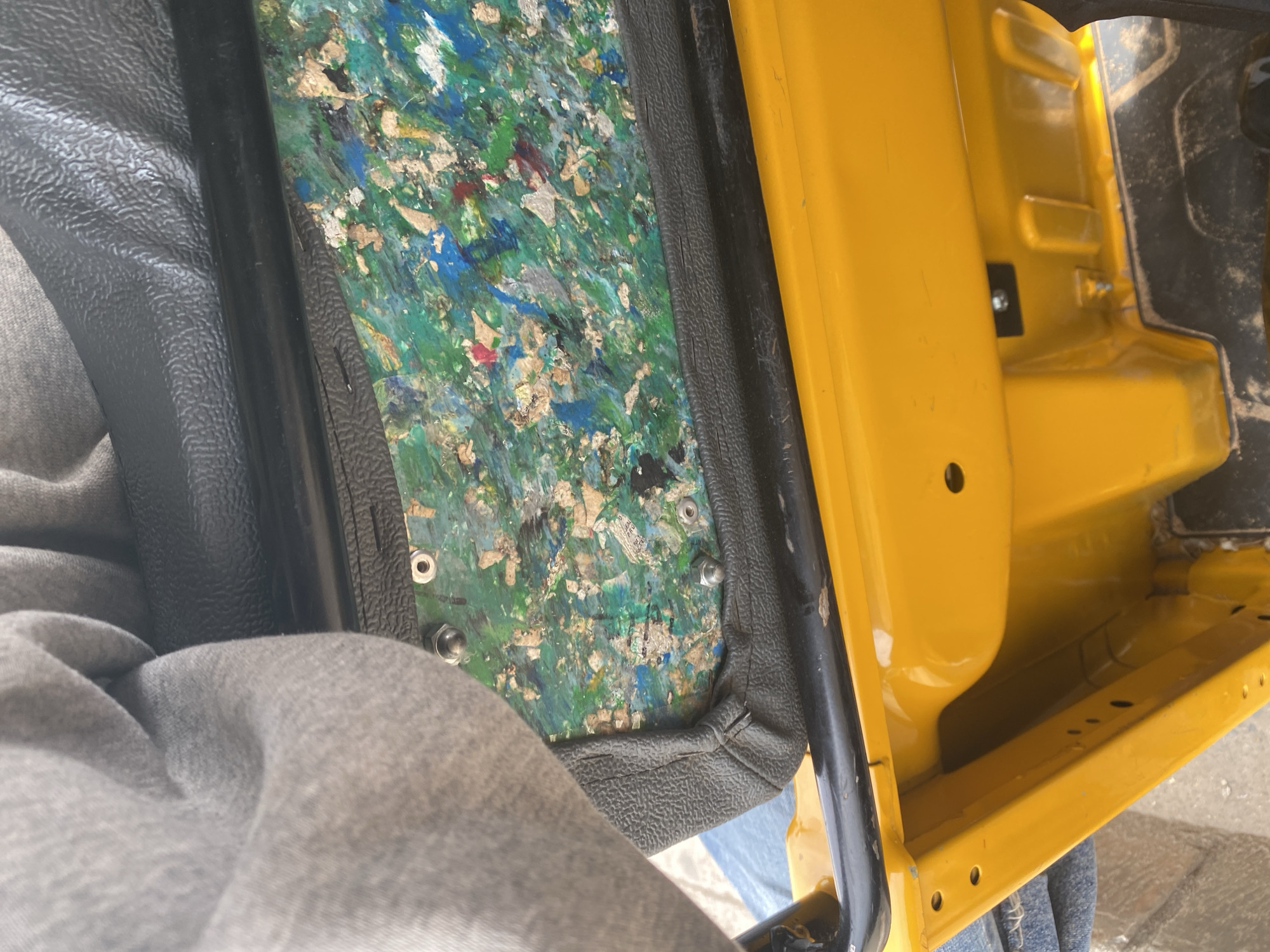The image captures a close-up view of a vehicle's interior, likely a tricycle or auto-rickshaw, in Jalingo, Nigeria. A grey, textured fabric, possibly clothing, is draped over a black, textured surface, which appears to be a seat or padding. Beneath this, a colorful, composite material is visible, featuring a mix of green, blue, white, and red fragments. Several small, metallic fasteners are embedded in this material. A bright yellow metal frame, part of the vehicle's body, is prominent in the lower portion of the image, showing some wear and tear. The overall scene suggests a utilitarian vehicle, possibly modified or undergoing repair, situated in an outdoor urban environment characteristic of Jalingo.