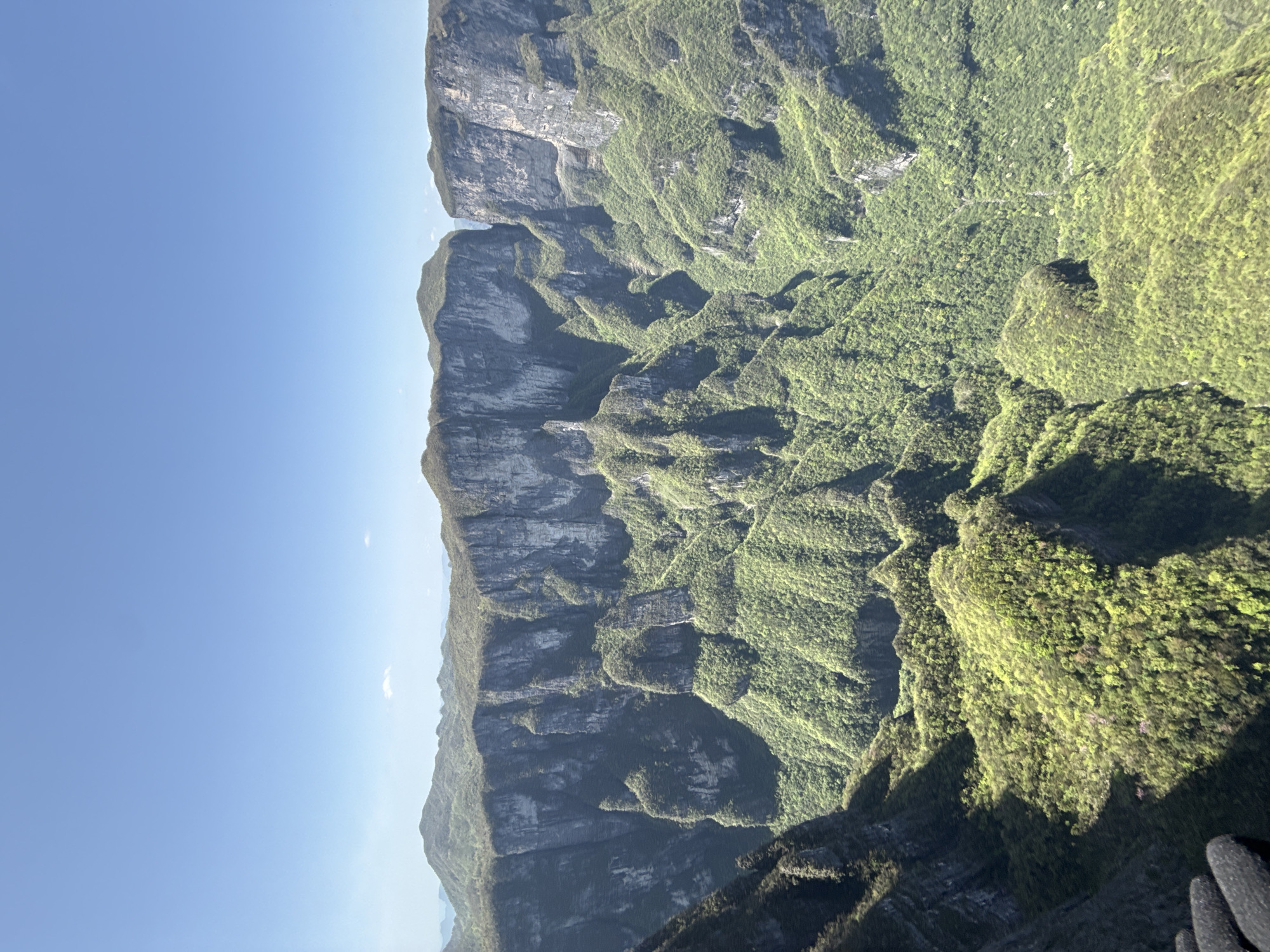 An eye-level, vertical shot captures a mountain range in Yongding District, China. The clear, light blue sky fills the left side. The landscape is characterized by steep, gray rock formations and lush, green vegetation that blankets the lower slopes and valleys. Shadows create depth and contrast on the terrain. No people are visible.