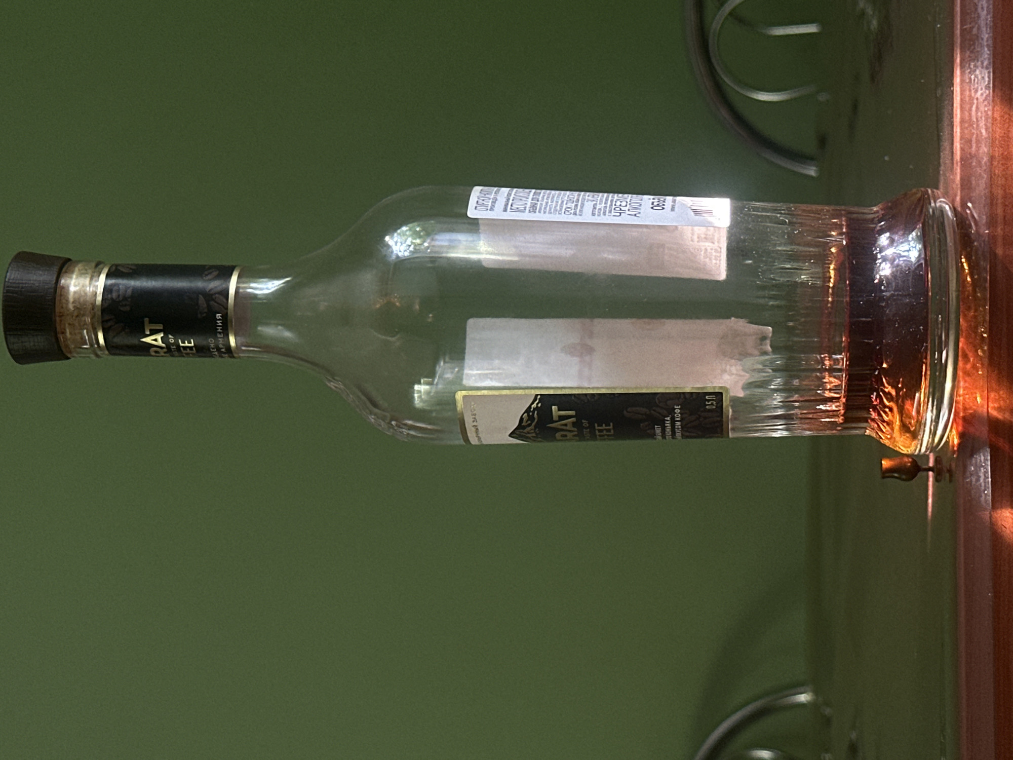 A close-up, eye-level shot focuses on a partially filled glass bottle of what appears to be brandy, labeled "ARARAT TASTE OF COFFEE." The bottle, standing on a dark, reflective surface, contains a dark amber liquid. Light refracts through the liquid and the ridged glass bottom, casting warm highlights on the surface. To the left of the bottle, a small, empty glass with a delicate stem sits on the surface, catching a glint of light. The background is a solid, dark green wall, creating a minimalist setting. Two curved metallic chair backs are visible in the upper right and left corners, suggesting an indoor setting, possibly a kitchen or dining area. The lighting appears to be from the right, casting long shadows. The scene does not provide any information that could place it in the city of Lobacheve, Ukraine.