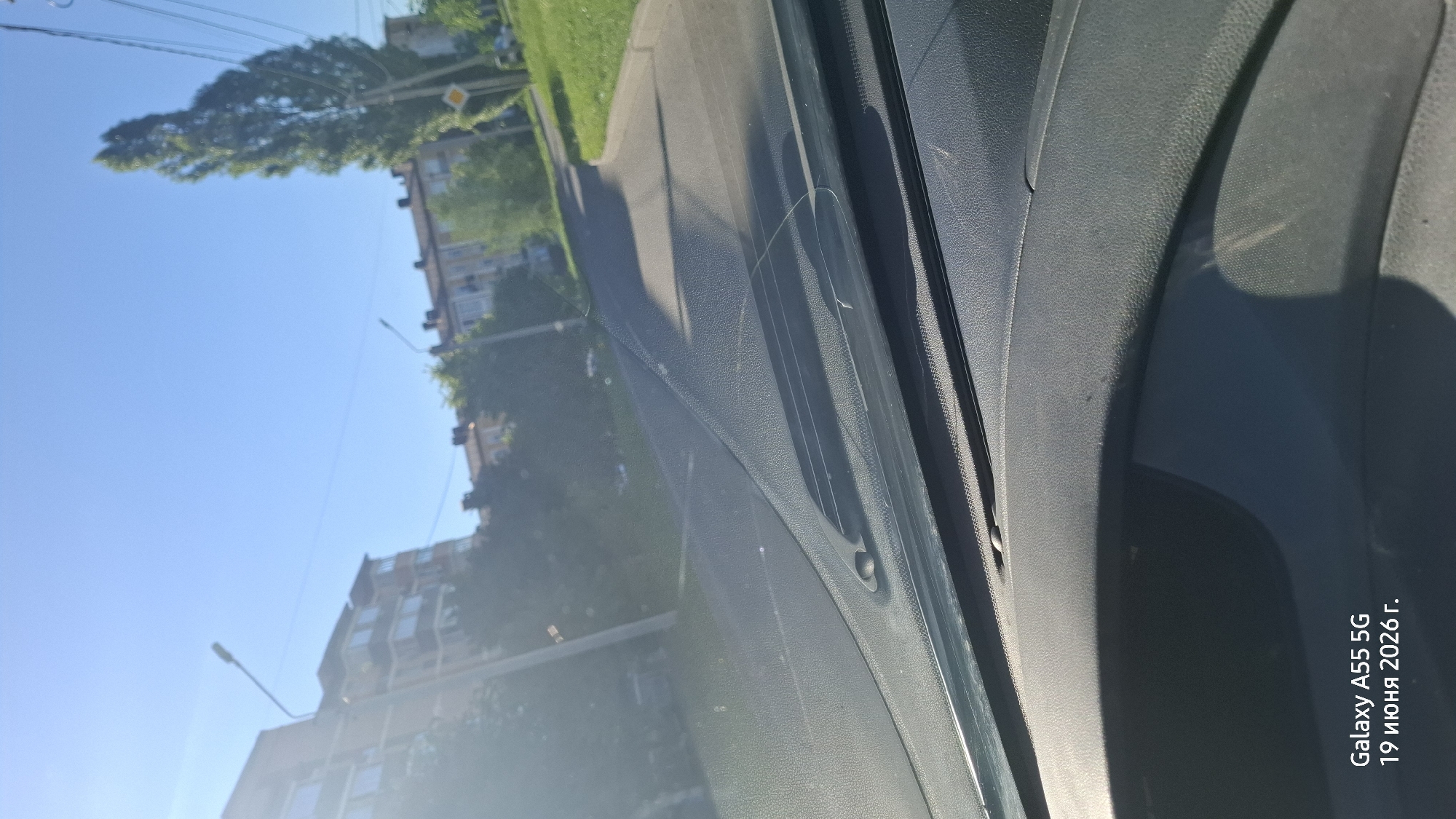 The image is an outdoor scene, likely taken from inside a vehicle, showing a view of a street and buildings. The sky is clear and blue, indicating a sunny day. There are trees with green foliage, suggesting it is daytime, likely morning or afternoon. The scene includes a paved road, a sidewalk, and multi-story buildings in the background. There is also a street lamp visible. The perspective suggests the photo was taken while moving, possibly in a car, as indicated by the interior elements in the foreground, such as a windshield or dashboard.
Visible text in the image reads: "Galaxy A55 5G 19 июня 2026 г." This indicates the device used to take the photo and a date, which appears to be in the future. Given the location context of Rostov-on-Don, Russia, the scene depicts a typical urban or suburban street view in that region. There are no people or specific events clearly depicted. The overall mood is calm and ordinary, reflecting a daytime street scene.