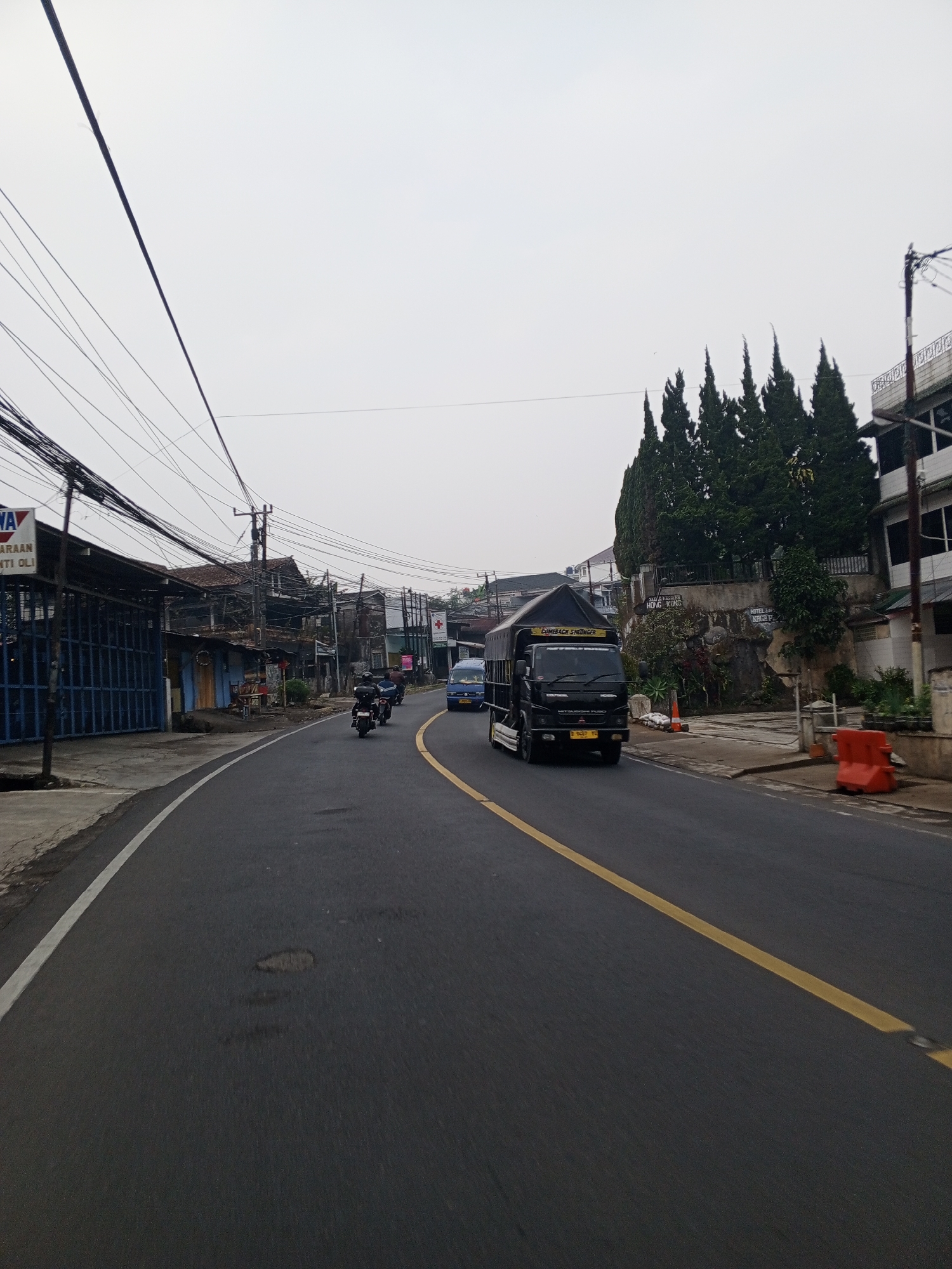 This is a street-level, eye-level photograph taken on an overcast day, looking down a paved road in what appears to be a town or city. The road has a white line on the left edge and a yellow double line in the center. In the foreground, the road surface shows some wear and tear, with a few potholes visible.
On the left side of the road, there is a row of buildings with a blue metal gate in front of the first structure, which has a sign with blue and white lettering. Further down the street, more buildings are visible, some with awnings and open fronts, with a significant amount of electrical wiring overhead.
In the middle of the road, a black truck with a "COMEBACK SHINE" sticker on its side is traveling away from the camera. To the left of the truck, two motorcycles are also moving in the same direction, with riders visible. Further ahead, a small blue van is also on the road.
On the right side of the road, a steep embankment is covered with dense green trees. Behind the trees, parts of buildings can be seen, including a white building with a decorative railing on its upper level. A sign with Chinese characters and the words "HOTEL BA" and "NEGA PA" is visible on the embankment. Several orange traffic barriers are placed along the side of the road.
The sky is a uniform light gray, indicating cloudy conditions. The overall impression is a busy street scene in a developing area, characterized by mixed architecture, commercial activity, and a tangle of overhead wires.