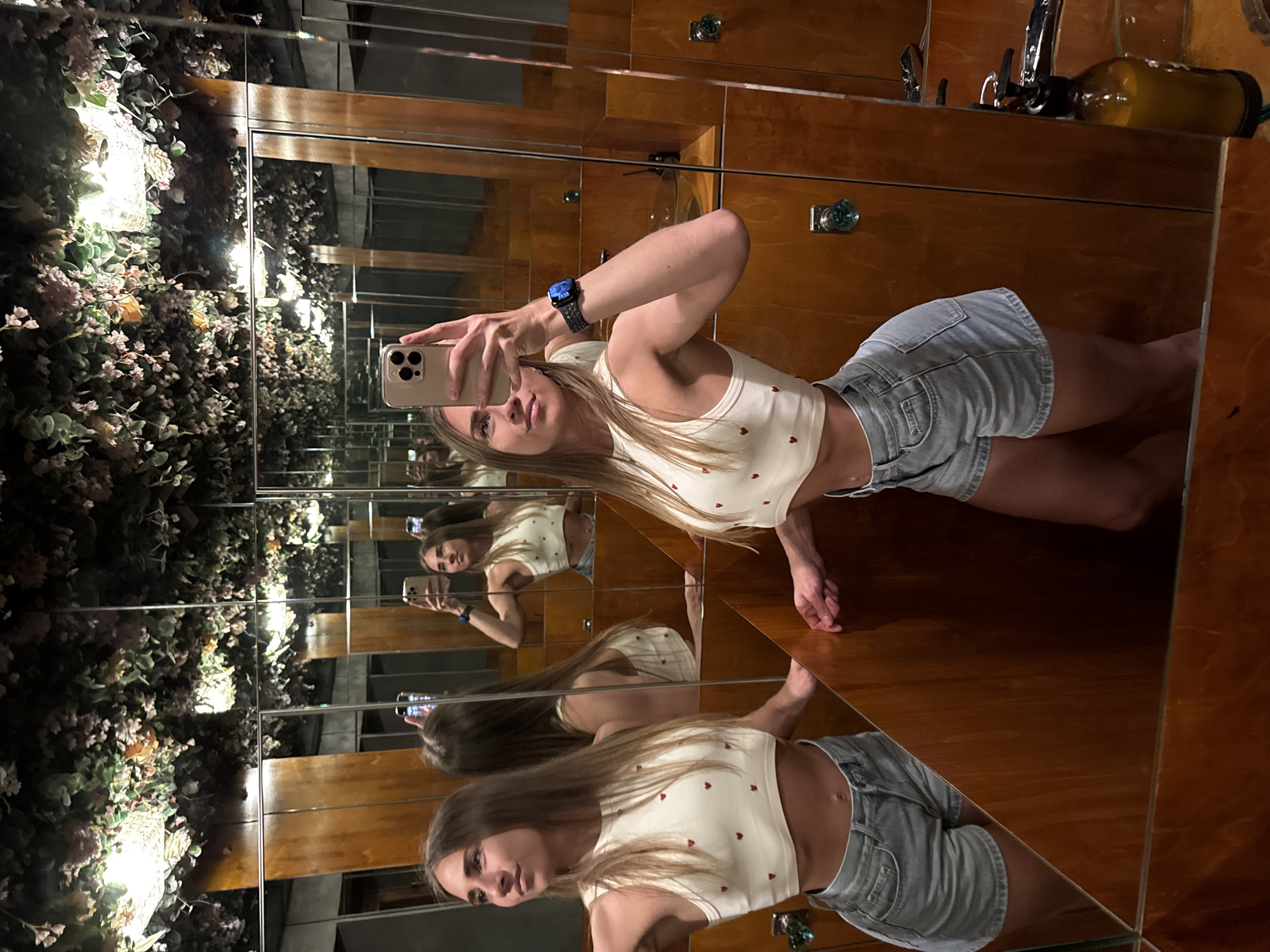 A young woman with long, brown hair is taking a selfie in a dimly lit restroom. She is wearing a white crop top adorned with small red hearts and light blue denim shorts. Her left arm is raised, holding a smartphone, and her right arm is bent at the elbow. A smartwatch with a dark band is visible on her right wrist. The restroom features mirrored walls that create multiple reflections of the woman and the space. The walls are paneled with dark wood, and decorative foliage hangs from the ceiling, interspersed with lighting fixtures. The overall impression is that of an intimate, possibly boutique, establishment. The location in Saint Petersburg, Russia, is not identifiable from the visual information presented in the image.