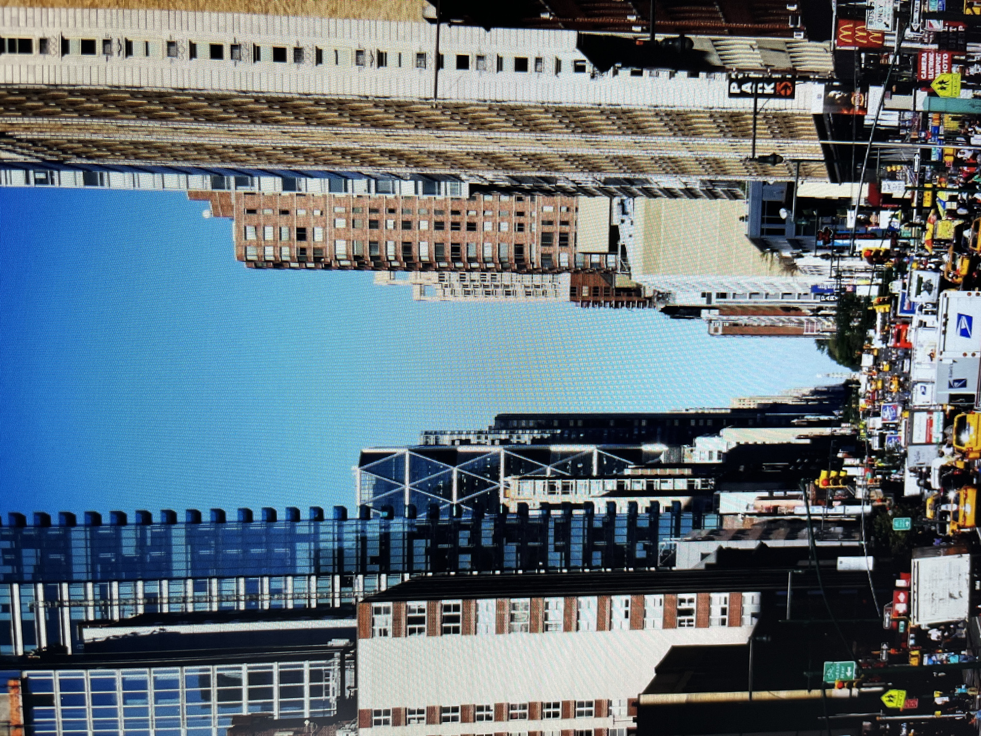 The image depicts a street-level view of a bustling city scene, likely in New York City given the presence of yellow taxis and tall, modern architecture. The perspective looks down a wide avenue flanked by towering buildings on both sides, their facades a mix of glass, steel, and brick. The sky above is a clear, vibrant blue.
In the foreground, the street is filled with traffic, dominated by numerous yellow taxis, white delivery trucks, and other vehicles. Pedestrians are visible on the sidewalks, some waiting at crosswalks. Traffic lights and street signs are abundant, indicating a busy intersection. The buildings vary in architectural style, from sleek, contemporary skyscrapers with geometric patterns to older, more traditional structures. Some buildings feature balconies and numerous windows.
Further down the avenue, the density of traffic and buildings continues, creating a sense of urban depth. The overall impression is one of a lively and dynamic urban environment on a bright, sunny day.