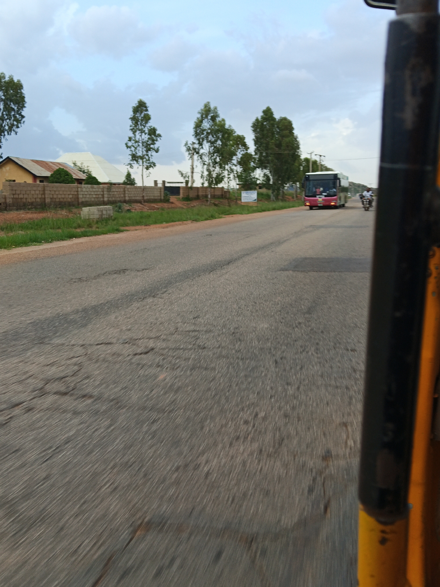 An eye-level, medium shot shows a street scene in a rural or suburban area under a cloudy sky. The foreground is dominated by the blurred asphalt of the road, suggesting movement. To the left, a low brick wall borders a grassy area and some scattered building materials, with a yellow building and a larger white-roofed structure visible behind it. Several trees are scattered along the roadside. In the distance, a red and white bus is traveling on the road, and a person on a motorcycle is also visible. The right side of the image is occupied by the blurred yellow and black frame of what appears to be a vehicle, possibly a tricycle or auto-rickshaw, from which the photo is taken.
