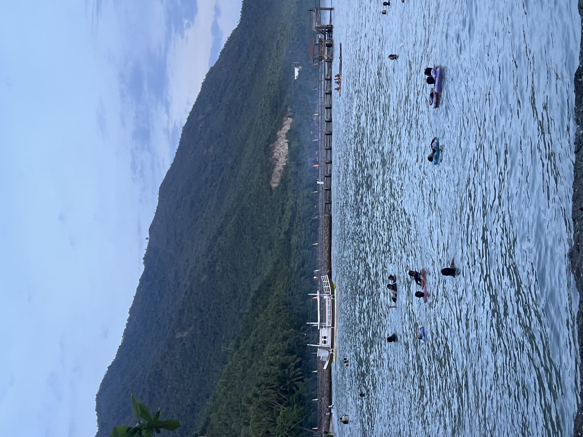 The image shows a beach scene in Banaybanay, Philippines. A lush, green mountain dominates the left side of the frame. A wooden pier stretches into the water from the base of the mountain. A white boat is docked at the pier. People are swimming in the water near the pier and the shoreline. The water is calm, with subtle ripples. The sky is overcast.