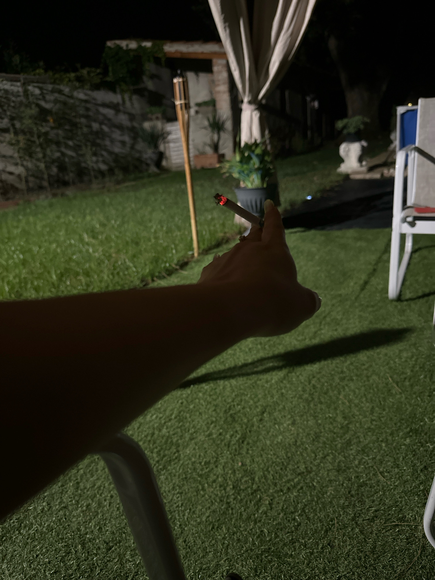 The image shows a close-up of a person's arm and hand holding a lit cigarette at night. The scene appears to be an outdoor patio or backyard, with artificial turf covering the ground. In the background, there is a tiki torch, a potted plant, and a white outdoor chair. The lighting is dim, suggesting it is nighttime. There is no indication of the location being Villa Pasquali, Italy.