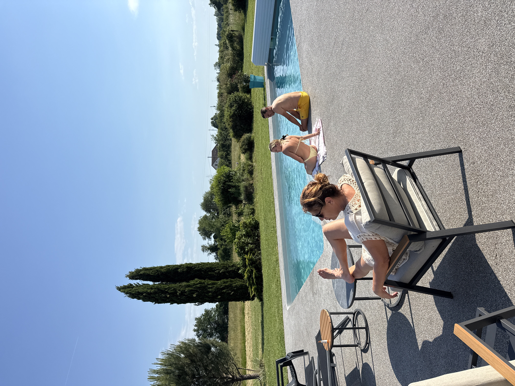 A sunny outdoor scene at a swimming pool in Veauchette, France. A woman sits in a modern chair near the pool's edge, placing her bare foot on a small round table. A man in yellow swim trunks and a woman in a bikini are near the pool. The surrounding environment includes trees, grass, a field, and a clear blue sky. The image is rotated counter-clockwise.