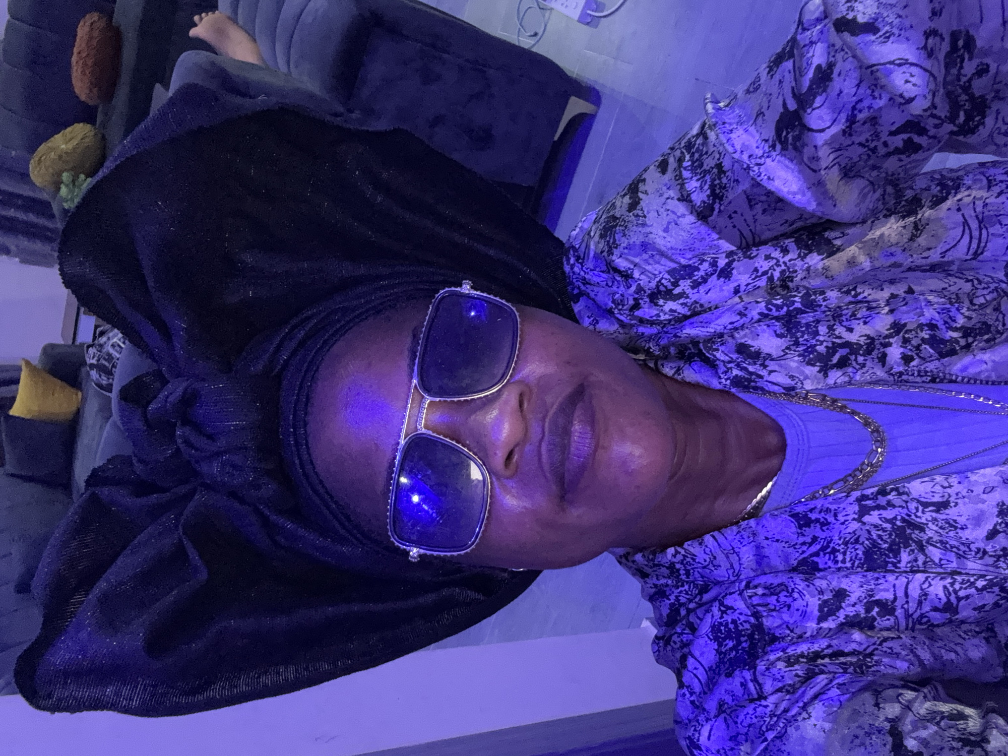A woman is photographed from the chest up, looking directly at the camera. She is wearing a large, black, sparkly head wrap styled in a prominent bow. She also has on large, square, silver-rimmed sunglasses with a reflective, blue-tinted lens. Underneath the head wrap, a darker fabric is visible, likely a scarf or the base of the head wrap. She is also wearing a patterned shirt with black and white abstract designs over a light blue top. Two gold necklaces are visible around her neck. The lighting in the scene is predominantly purple, casting a strong hue over the woman and her surroundings. In the background, a plush, dark blue couch is visible, with a person's bare foot resting on it. The floor appears to be tiled and is also reflecting the purple light. A portion of a couch with throw pillows in yellow and brown is also visible further in the background. The overall impression is a selfie taken in an indoor, domestic setting, possibly a living room.
