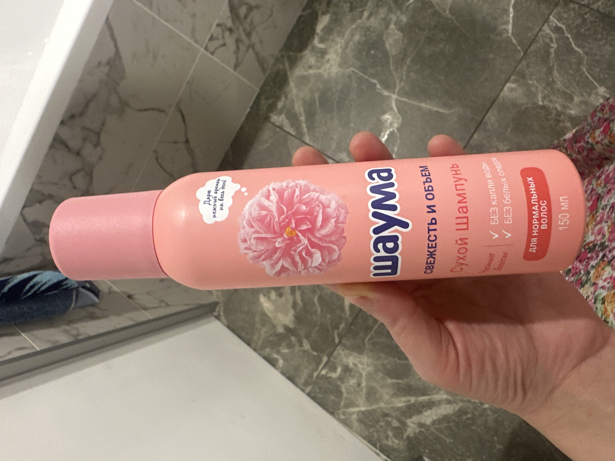 An overhead shot shows a person's hand holding a can of Syoss dry shampoo. The can is pink with a graphic of a pink flower and text in Russian. The background is a bathroom with gray marble tiles and a white bathtub.
