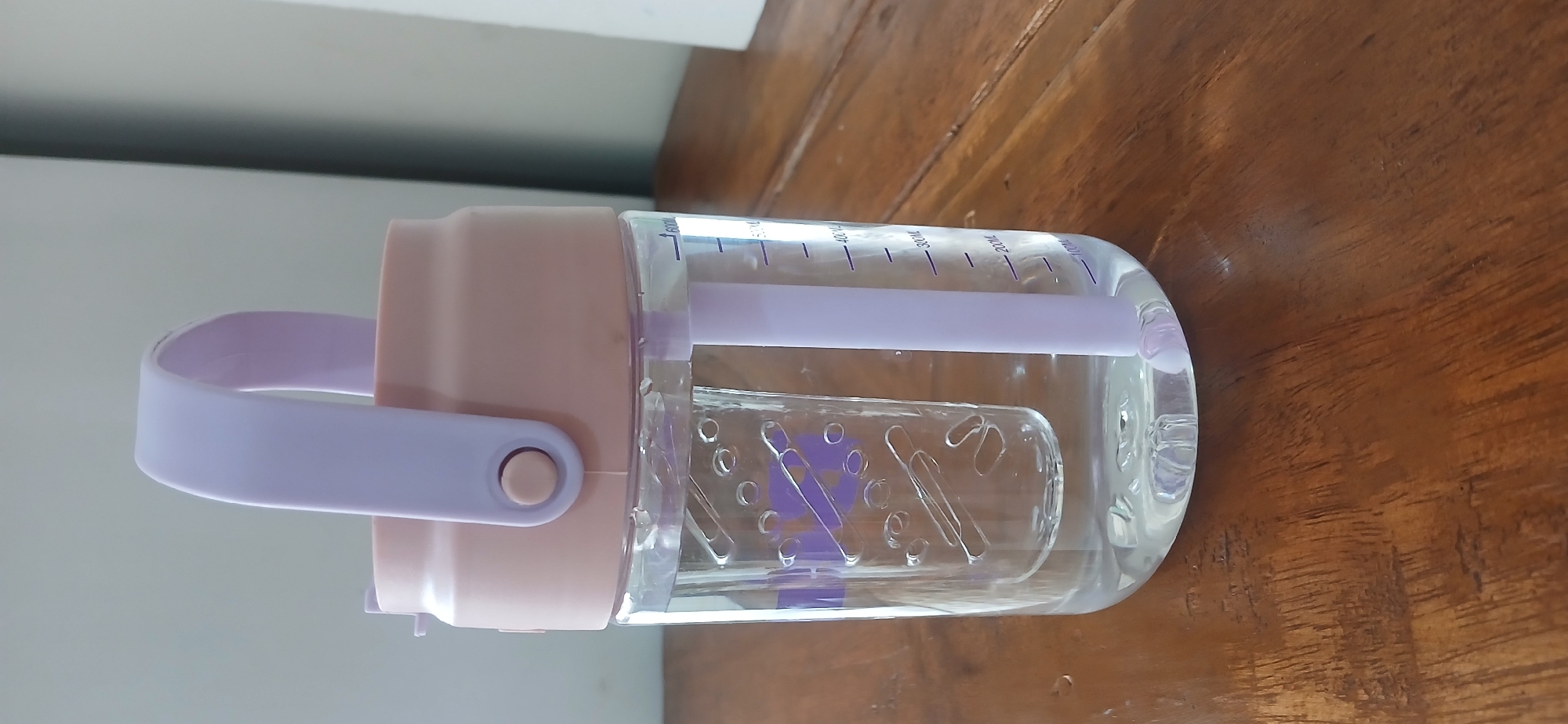 A clear plastic water bottle with a light pink lid and a lavender handle and straw is standing on a wooden table. The bottle contains clear liquid and has measurement markings on the side, indicating it holds up to 600ml. Inside the bottle, there is a clear plastic insert with a decorative pattern of circles and lines. The water bottle is positioned in the foreground, with the wooden table surface taking up the bottom portion of the image. The background is a plain, light-colored wall, suggesting an indoor setting. There are no people or animals present in the image. The lighting appears to be natural or from an overhead source, casting subtle shadows on the table. The image was taken in the city of West Jakarta, Indonesia.