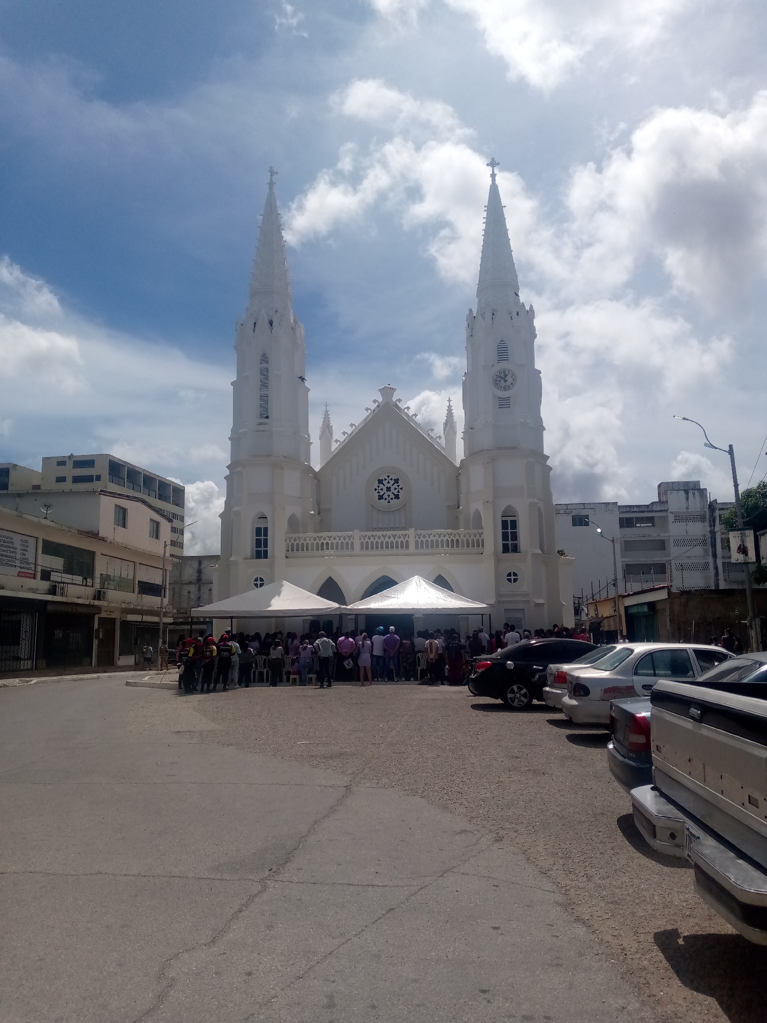 The image shows the Iglesia San Juan Evangelista in Juan Griego, Venezuela. The white church with two tall spires dominates the scene, with a small crowd gathered under a white tent in front of the entrance. Several cars are parked nearby. The road in front of the church is cracked and slightly damaged. Buildings of varying heights flank the church on either side. The sky is partly cloudy, with patches of blue visible.