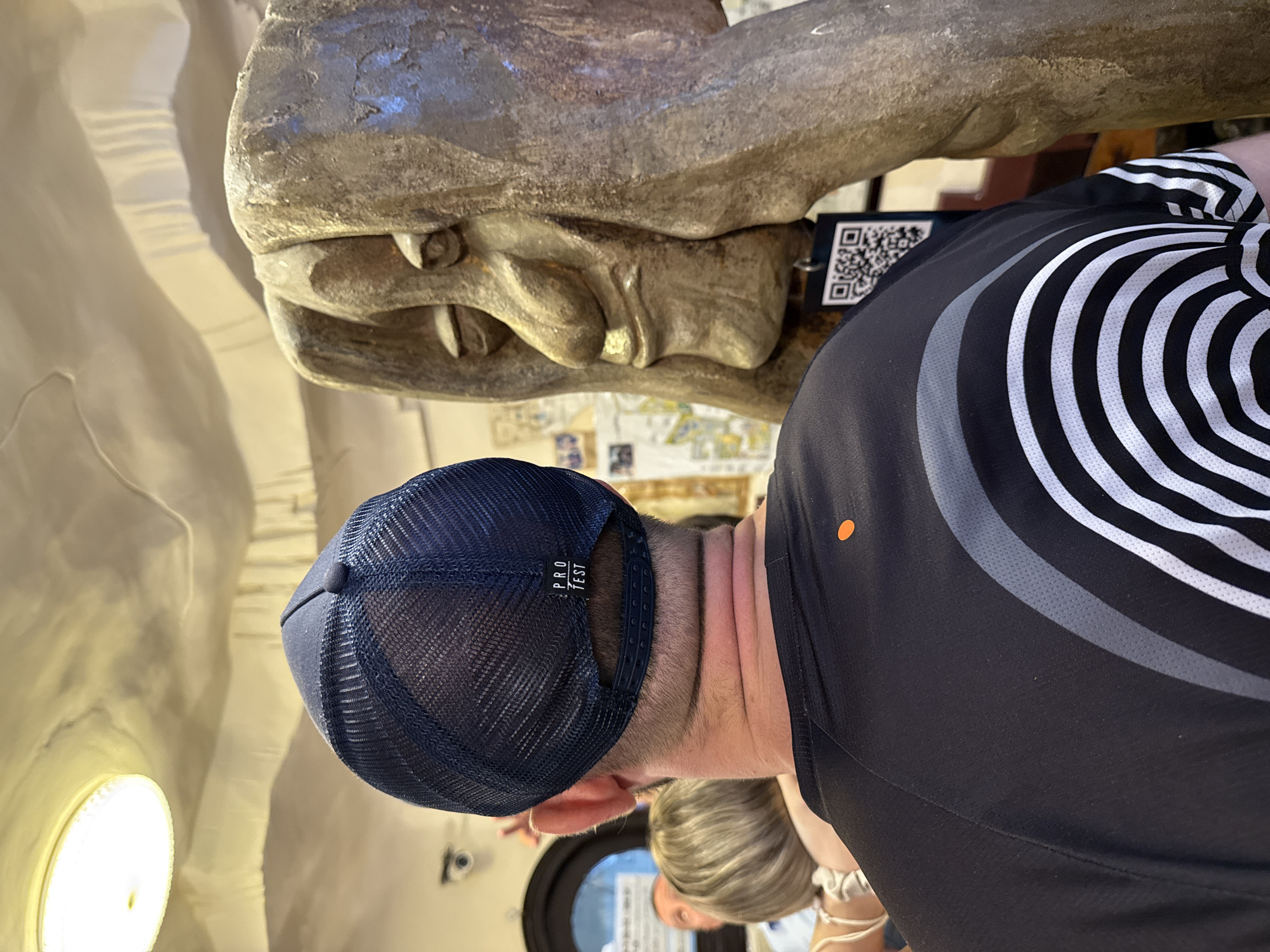 This vertical image, captured in Da Lat, Vietnam, depicts a person viewed from behind, standing before a large, weathered statue or sculpture. The individual wears a navy blue mesh baseball cap with "PRO TEST" visible on the back strap, and a black short-sleeved shirt featuring a distinctive design of black, gray, and white wavy stripes on the right side, along with a small orange dot on the upper left back.
The prominent statue, appearing to be made of stone or a similar material, showcases a large head or part of an animal/mythical creature with visible, pointed teeth or tusks. A black tag bearing a QR code hangs from the statue, likely offering information about the exhibit. The setting is indoors, possibly a museum or gallery, indicated by the statue and QR code, with light-colored walls and a circular light fixture visible on the left. Another person's blonde hair is partially visible in the lower background. The scene suggests an observation or appreciation of art or history.