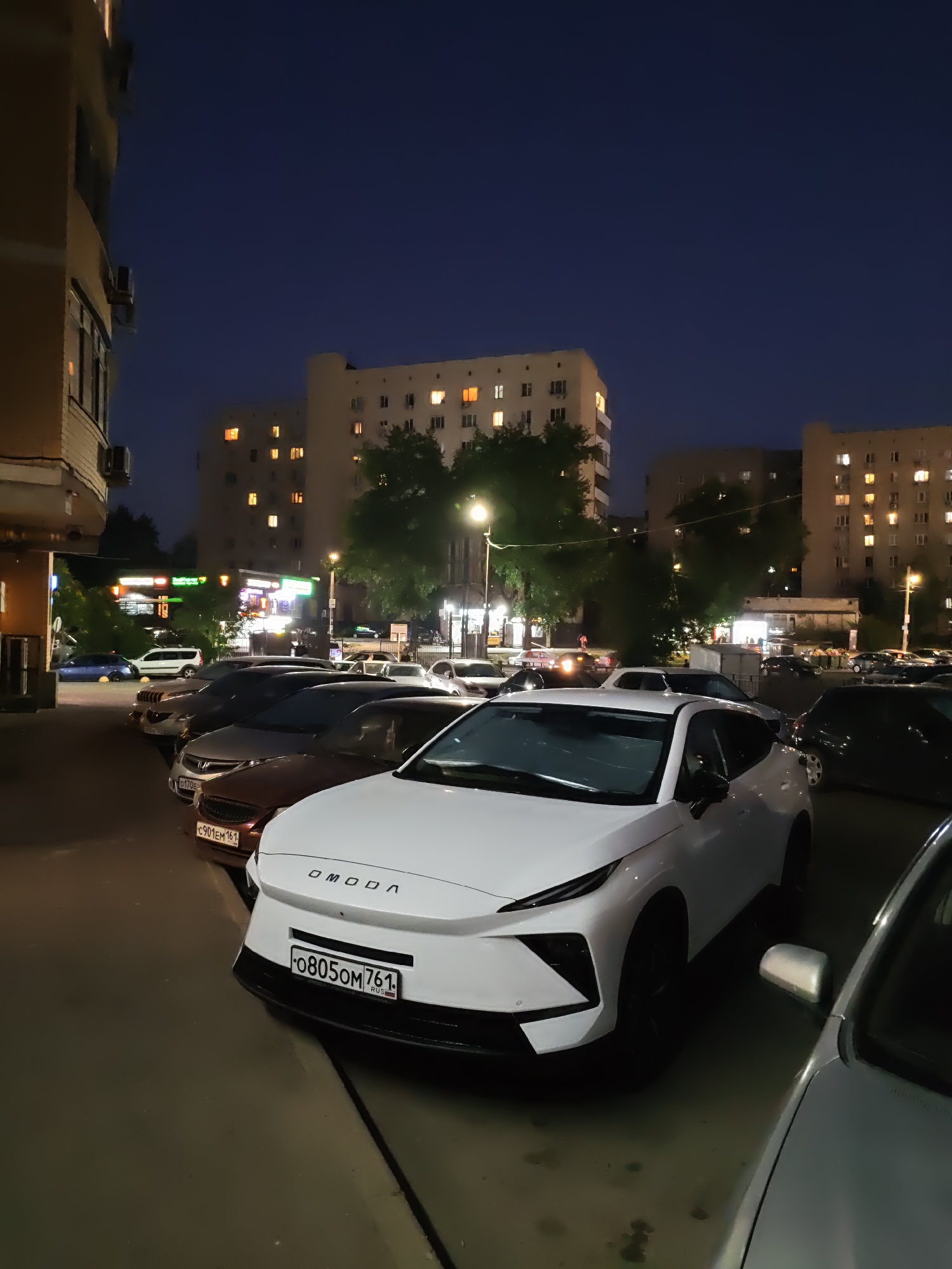 The image depicts a street scene at night in Rostov-on-Don, Russia. The foreground is dominated by the front of a white OMODA car, license plate "F0805OM 761 RUS". To the left of the OMODA, there is a row of parked cars, including a dark red car with license plate "C901EM 161 RUS". In the background, there are several multi-story apartment buildings with illuminated windows. Trees are visible between the buildings and the street. Streetlights and the lights from shops are also present, creating a moderate level of illumination. The sky is dark blue, indicating nighttime. No people are visible in the frame.