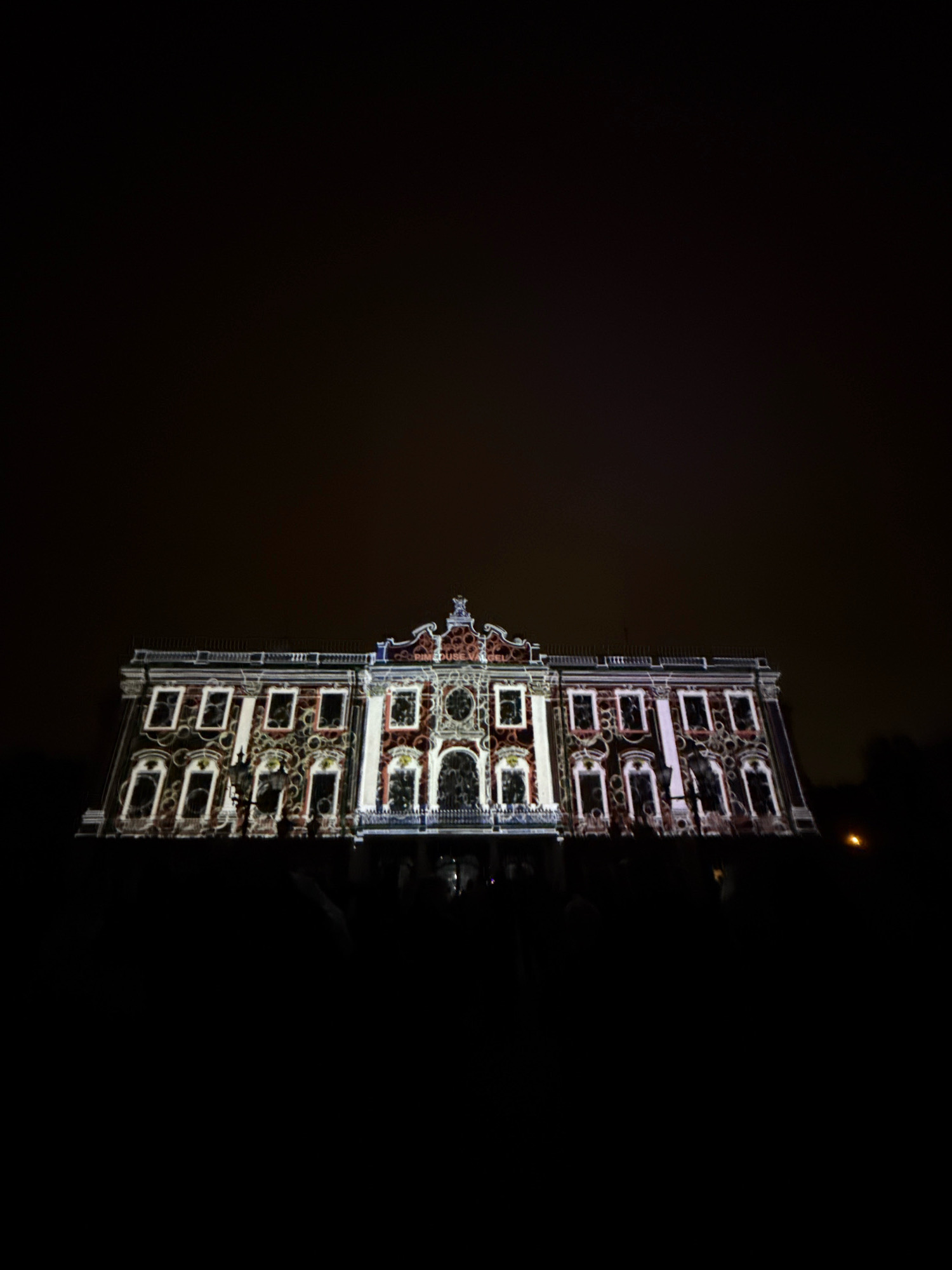 The image depicts a projection mapping light show on a building in Tallinn, Estonia. The building, likely a historical structure, is illuminated with complex patterns and designs, creating a dynamic visual display against the dark night sky. The projection covers the facade of the building, highlighting architectural details and creating an illusion of movement and depth. The lower part of the image is mostly black, indicating the presence of darkness and possibly a crowd gathered to watch the show. The overall scene suggests a festive or cultural event taking place in the city.