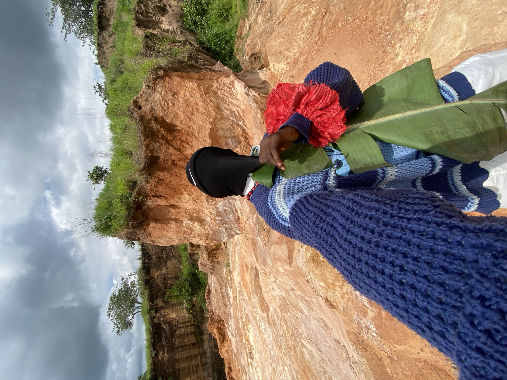 A person wearing a black head covering and a blue knit sweater with blue and white wavy patterns stands in a landscape with eroded orange earth and patches of green grass. The person holds a red flower-like object and two green banana leaves. In the background, there are steep, layered orange cliffs, vegetation, and a cloudy sky. The ground appears to be composed of loose soil and small rocks.