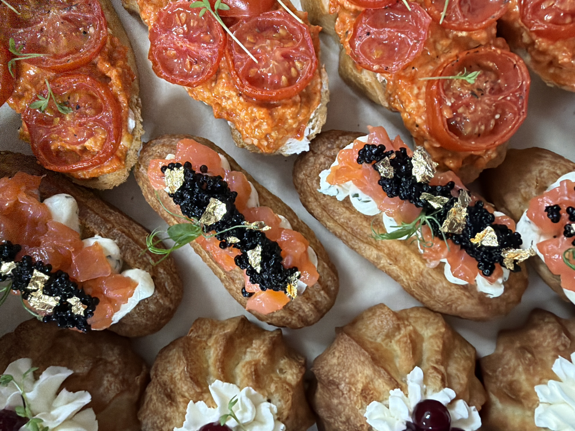 An overhead view displays an assortment of appetizers arranged on a light-colored surface in Chaikovsky, Russia. The scene features three distinct types of culinary preparations. On the left side of the frame, several golden-brown choux pastry items are topped with a swirl of white cream, each garnished with a single dark red berry and a sprig of green microgreens. Centrally, three elongated choux pastries are dressed with a white creamy base, layered with pieces of salmon, topped with black caviar, interspersed with gold leaf flakes, and adorned with green microgreens. On the right side, multiple oblong pieces of toasted bread are covered with an orange-red spread, layered with thin slices of red tomato, and garnished with green microgreens. No individuals are present. The items are closely arranged, filling the frame.