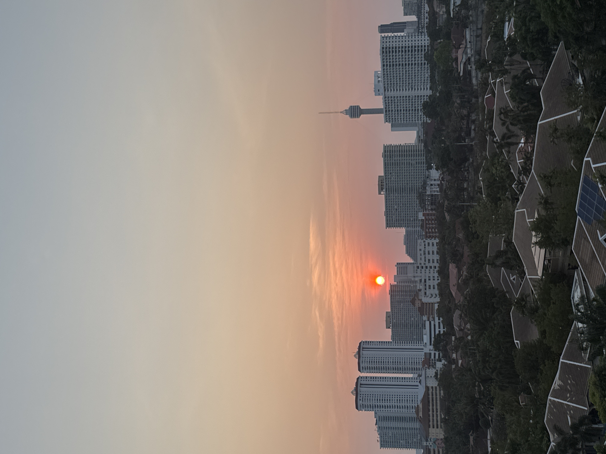 The image shows a cityscape at sunset in Pattaya City, Thailand. The sun, a bright orange disk, is setting behind a line of high-rise buildings. A tall communications tower is visible amidst the buildings. The sky is gradient, transitioning from a light gray-blue at the top to a warm orange near the horizon. Residential buildings with pointed roofs and solar panels are interspersed with green trees.