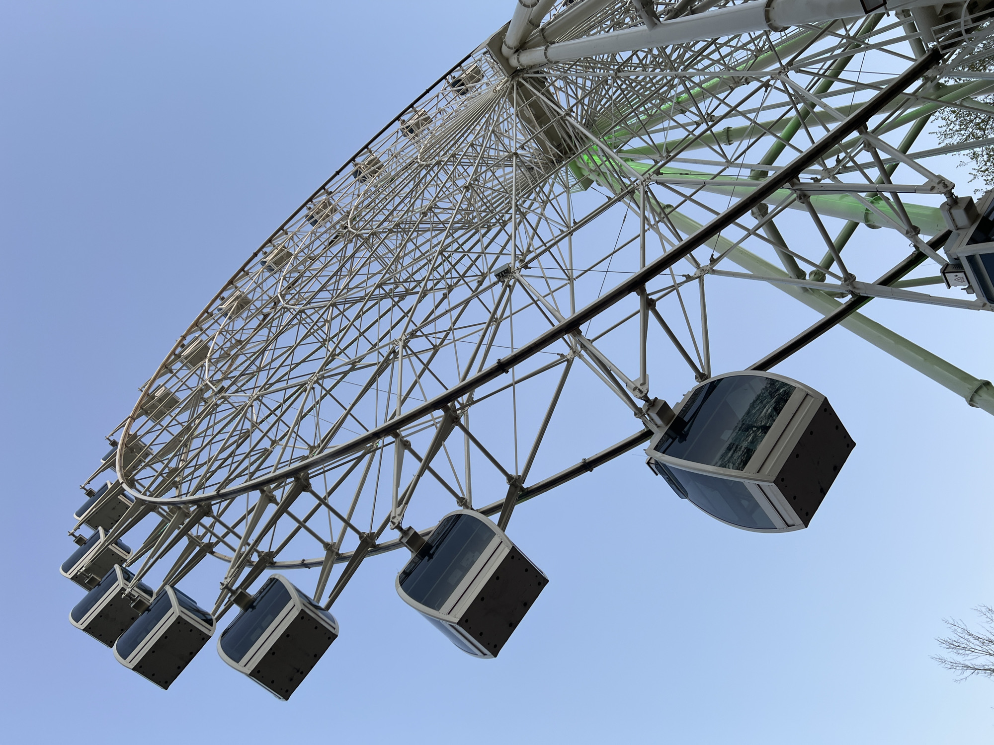 The image is a low-angle shot of a Ferris wheel in Cherepovets, Russia. The wheel structure is primarily white with gray gondolas. The gondolas are suspended from the wheel's outer perimeter. The sky is a clear, light blue. There are green lights along the wheel's support structure. Sparse tree branches are visible in the top right and bottom right corners.